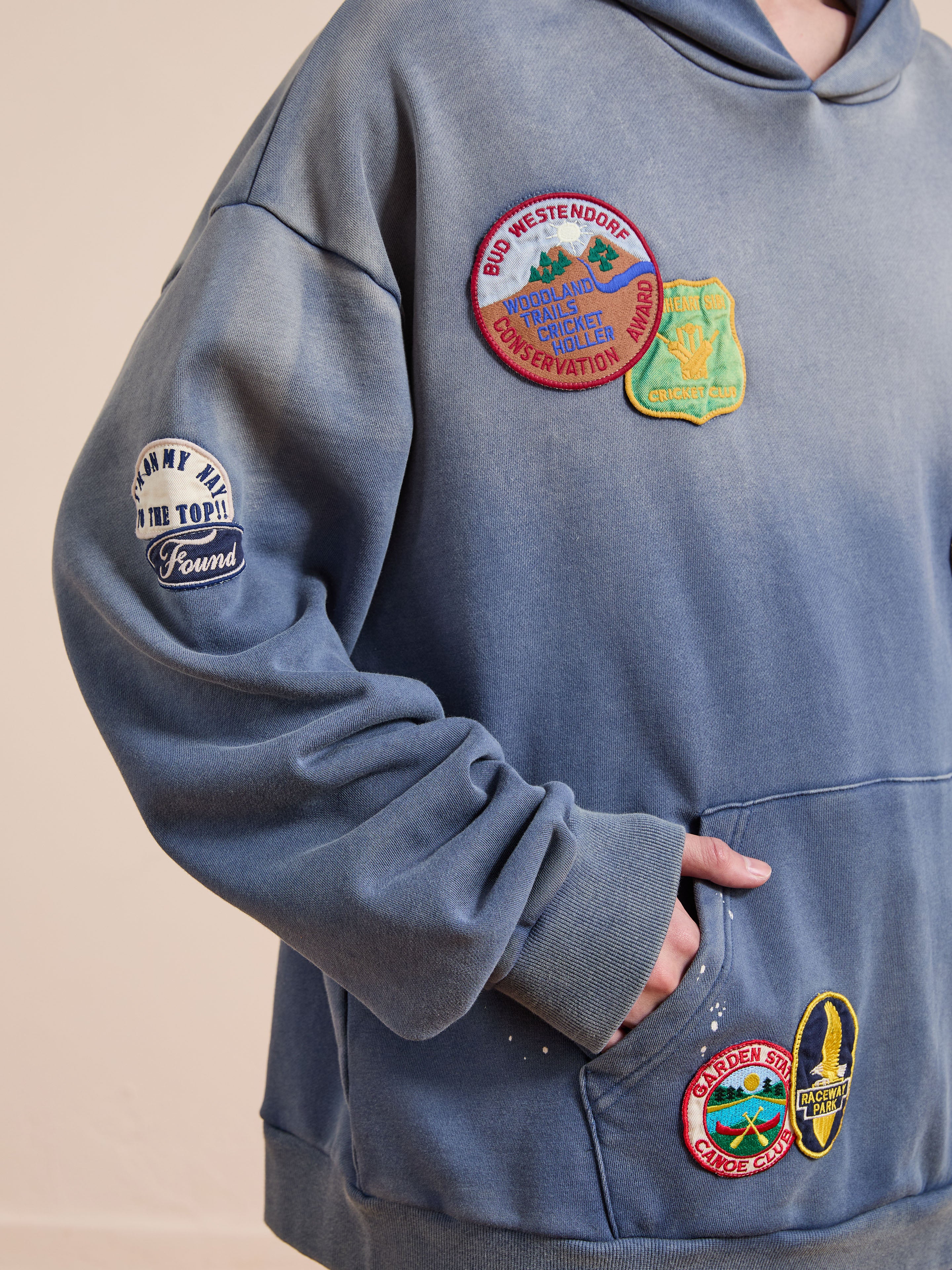A person wears the FOUND Distressed Patch Hoodie, featuring a blue vintage look with colorful embroidered patches—some with text and graphics—and stands with one hand in their pocket.