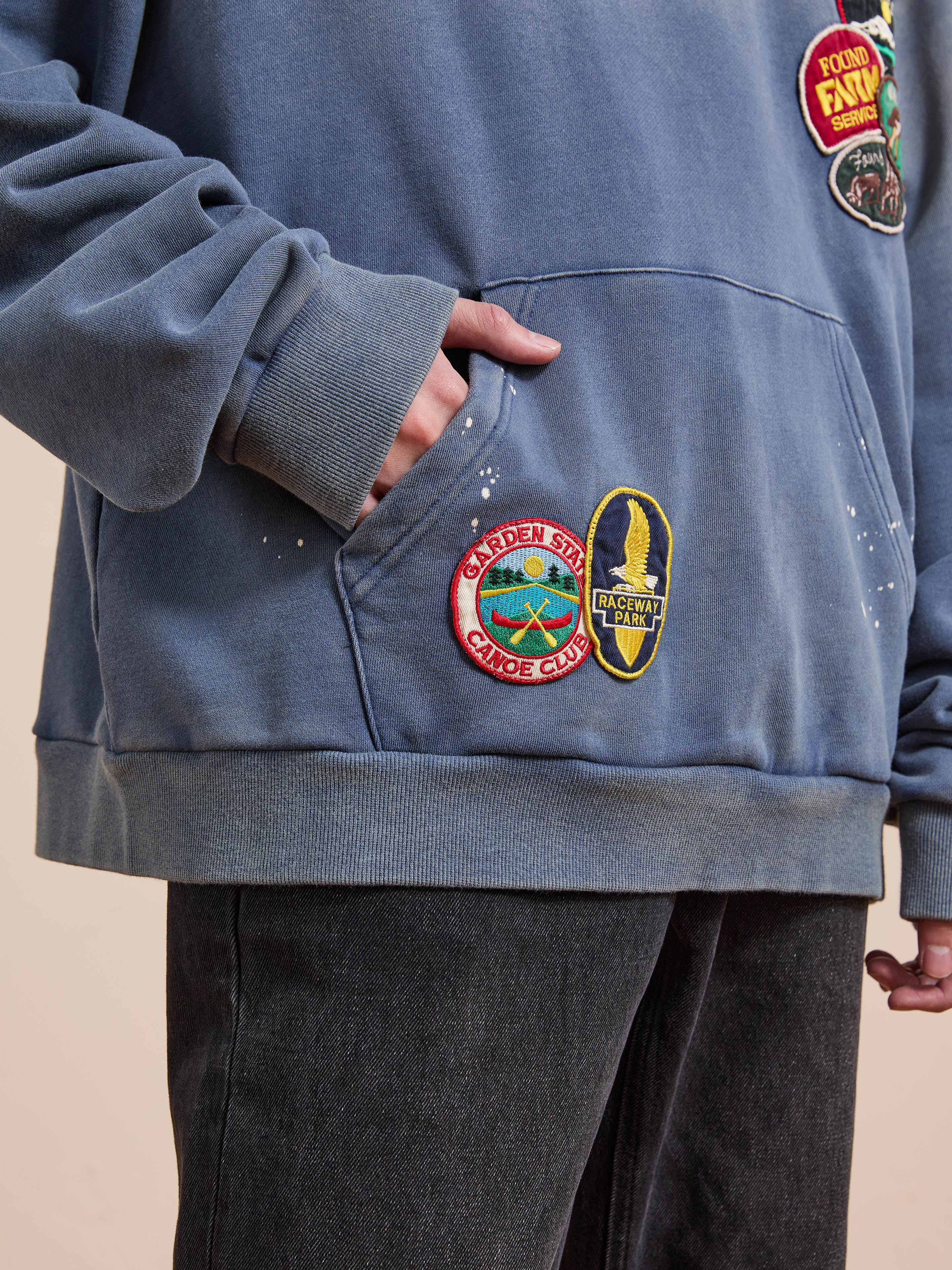 A person wearing the FOUND Distressed Patch Hoodie with colorful embroidery and black jeans, one hand tucked in the hoodie pocket.