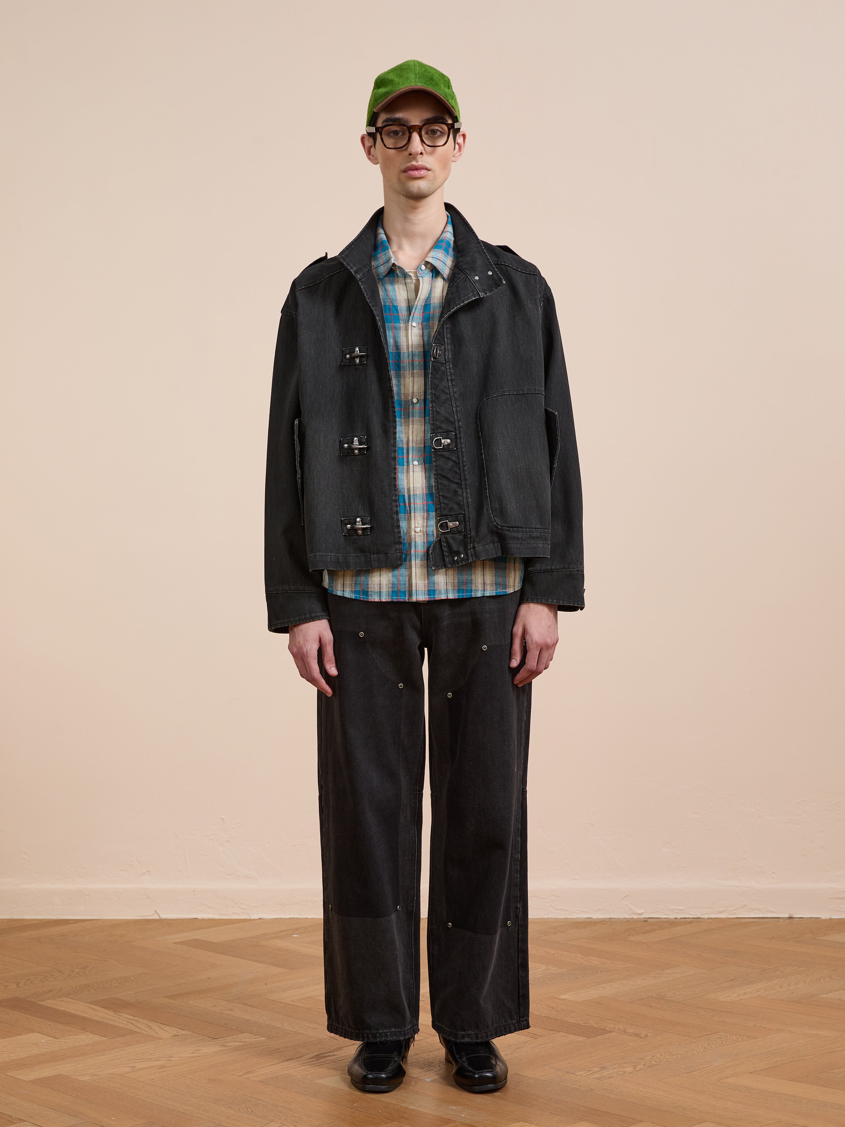 Indoors, a person wears the Found Utility Clasp Work Jacket over a plaid shirt, paired with a green cap, glasses, loose black pants, and black shoes on a wooden floor.