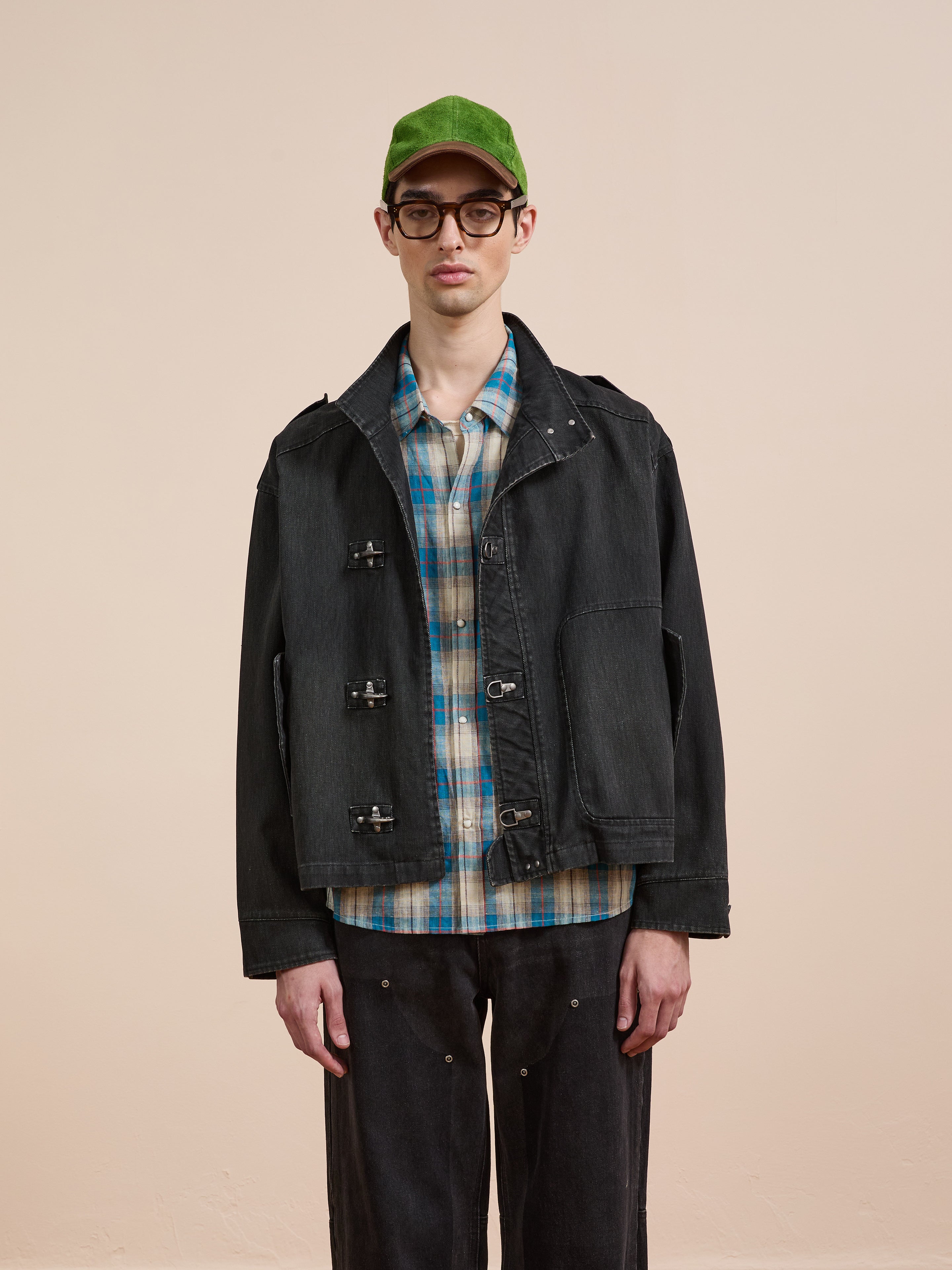 A person wears glasses, a green cap, plaid shirt, and dark pants with the Found Utility Clasp Work Jacket, standing against a plain beige background.