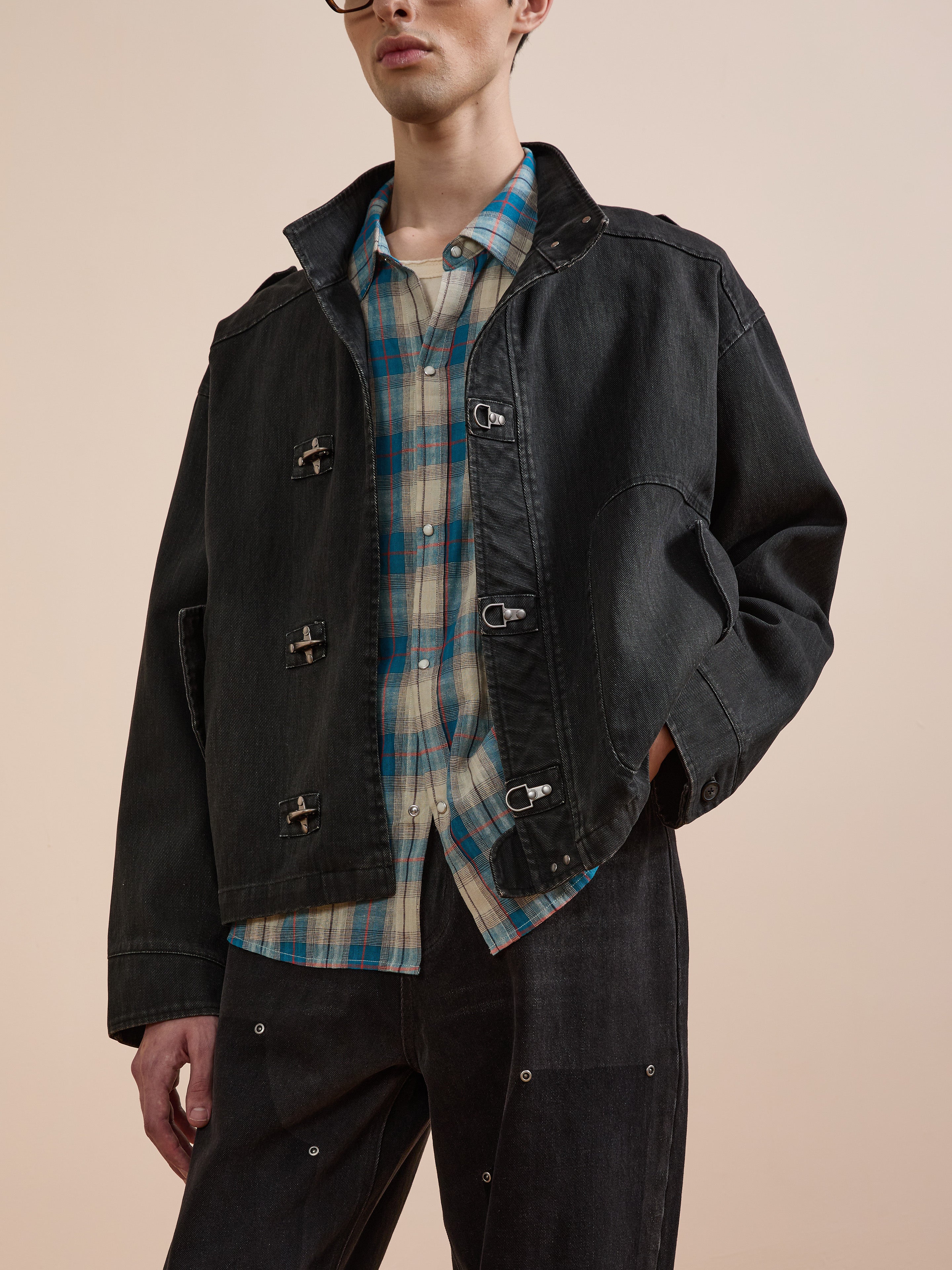 A person wears the Found Utility Clasp Work Jacket in vintage black over a blue and white plaid shirt, paired with black pants, standing against a neutral background.
