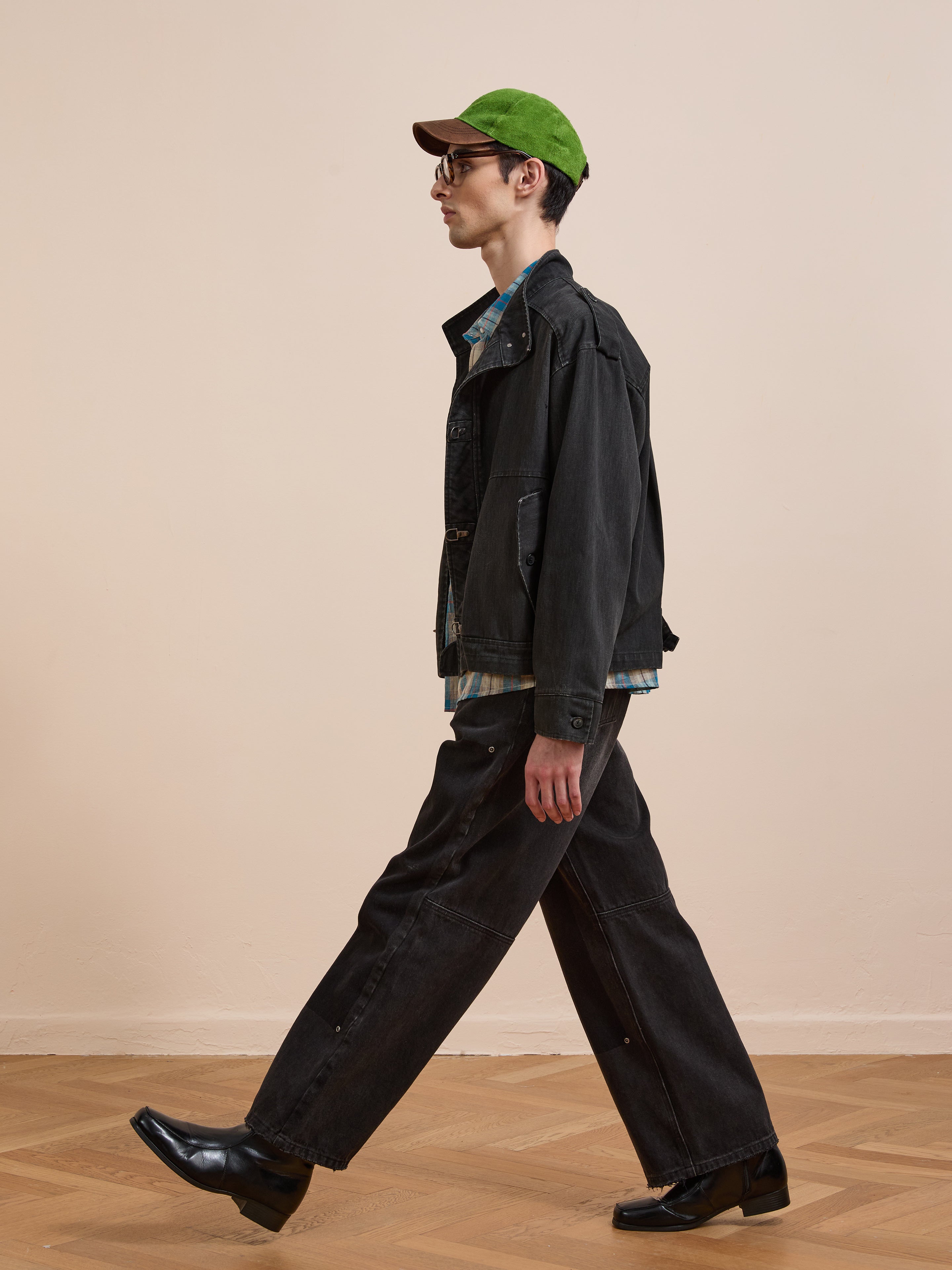 Wearing the Found Utility Clasp Work Jacket, black pants, green cap, sunglasses, plaid shirt, and black shoes, a person walks across a wooden floor with a plain beige backdrop.