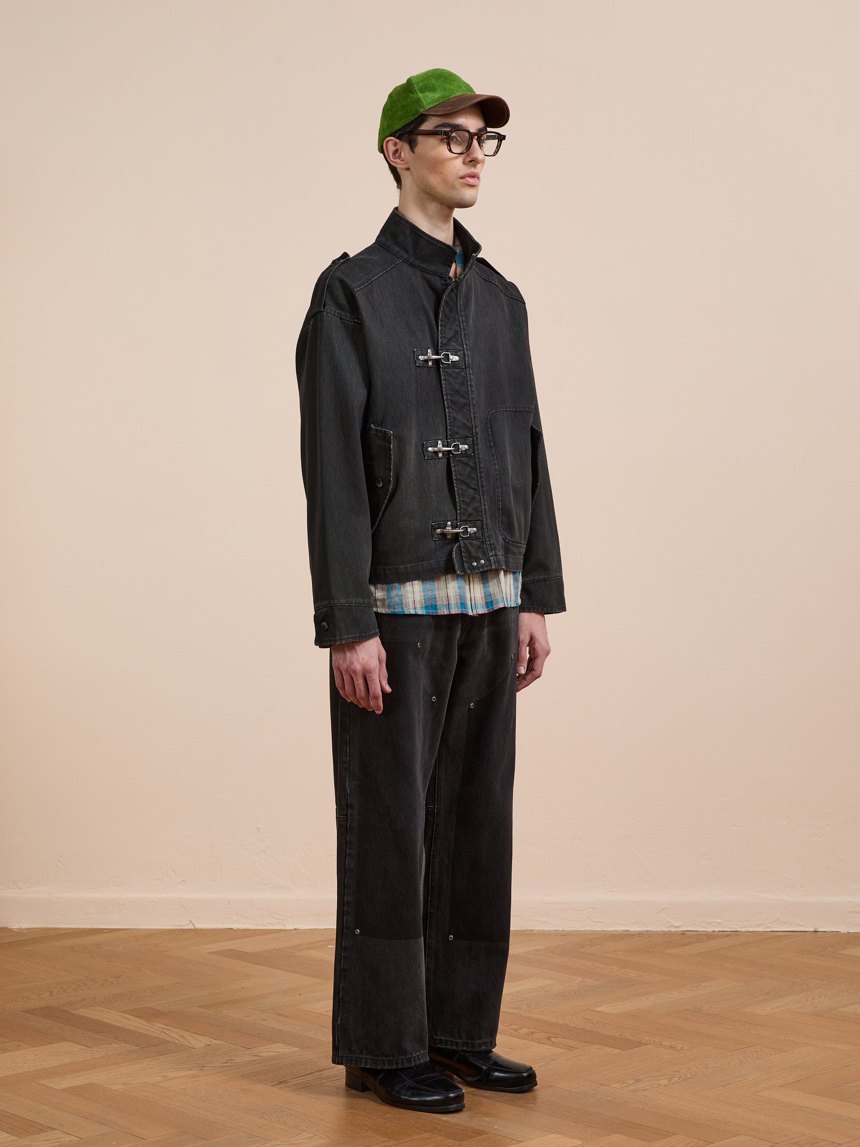 Indoors against a beige wall and wooden floor, a person wears a green cap, black-rimmed glasses, wide black pants, black shoes, and the Found Utility Clasp Work Jacket over a plaid shirt.