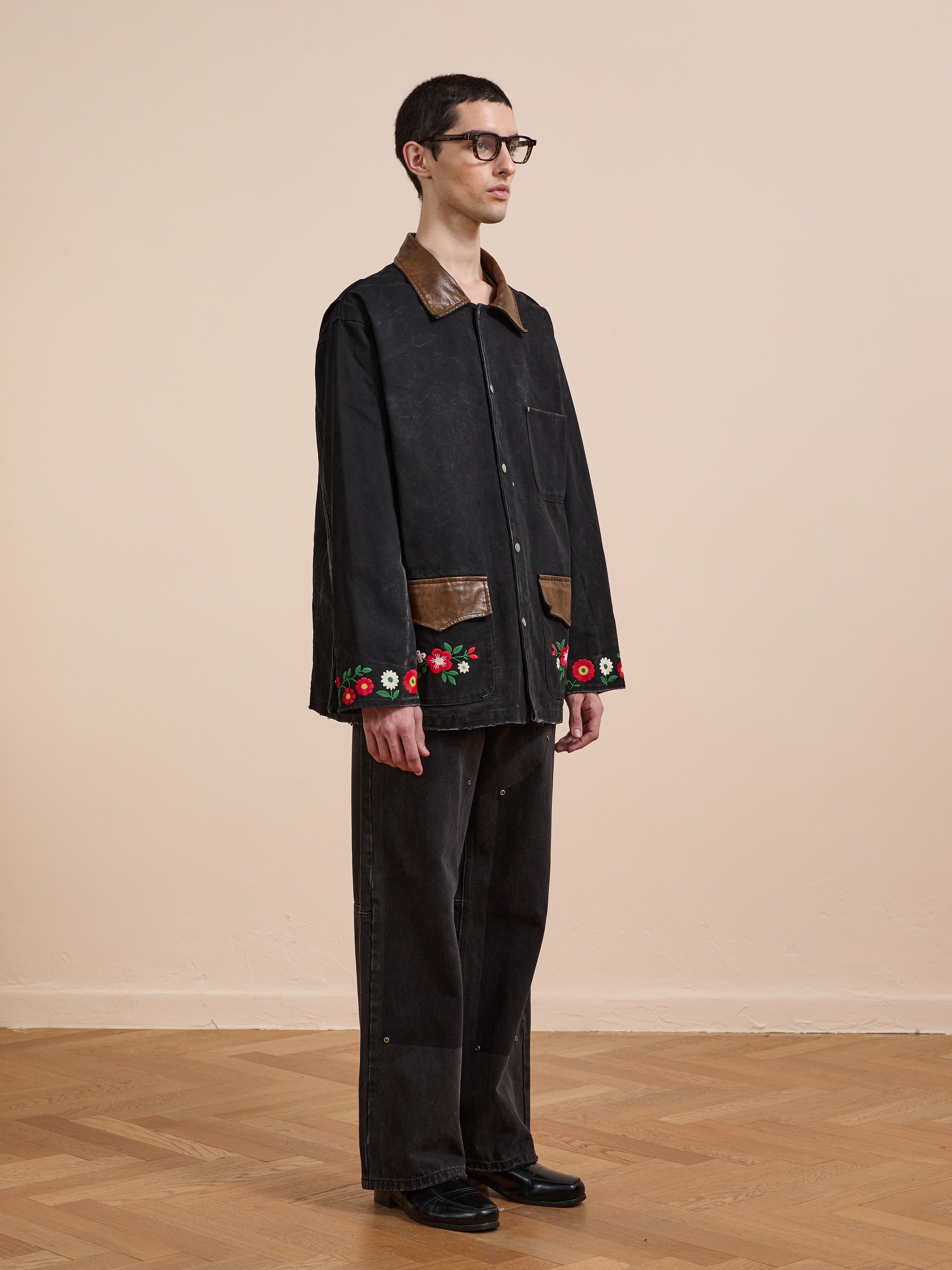 A person stands indoors wearing the Found Canvas Embroidered Work Jacket, featuring floral embroidery on the sleeves and brown leather trim, paired with black pants and black shoes.