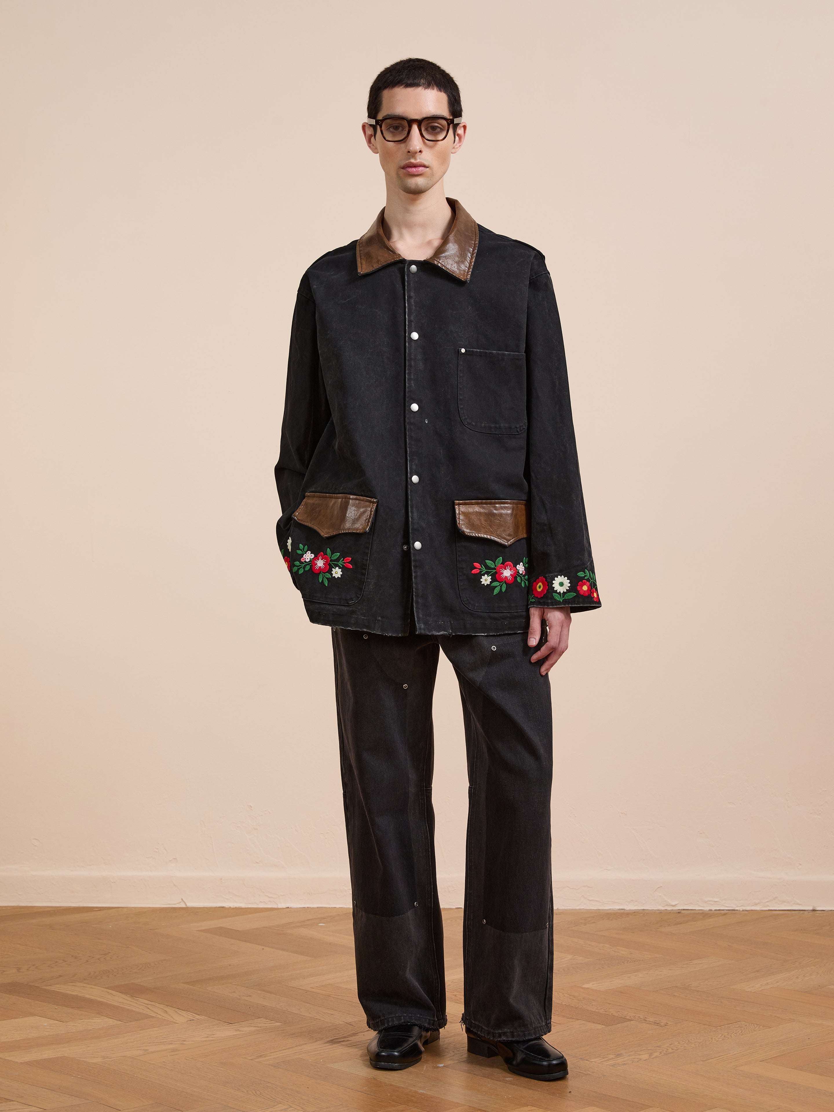 Indoors, a person wears the Found Canvas Embroidered Work Jacket featuring brown accents and floral embroidery, paired with unisex relaxed fit black pants, black shoes, and glasses. The background is plain with a wooden floor.