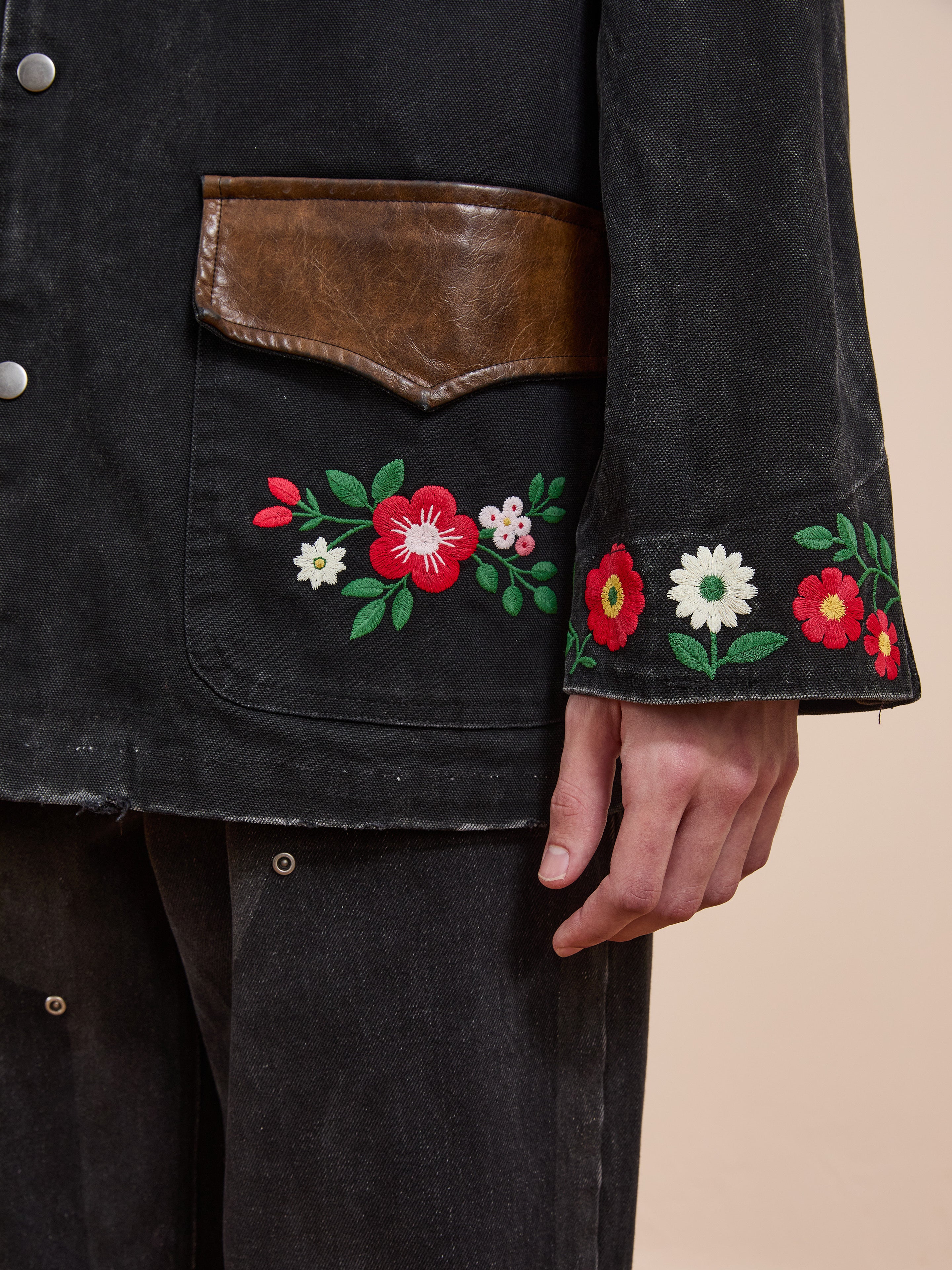 Someone wearing the Found Canvas Embroidered Work Jacket—a unisex, relaxed fit black canvas jacket with brown leather pocket trim and colorful floral embroidery on the sleeve and pocket—paired with black pants.
