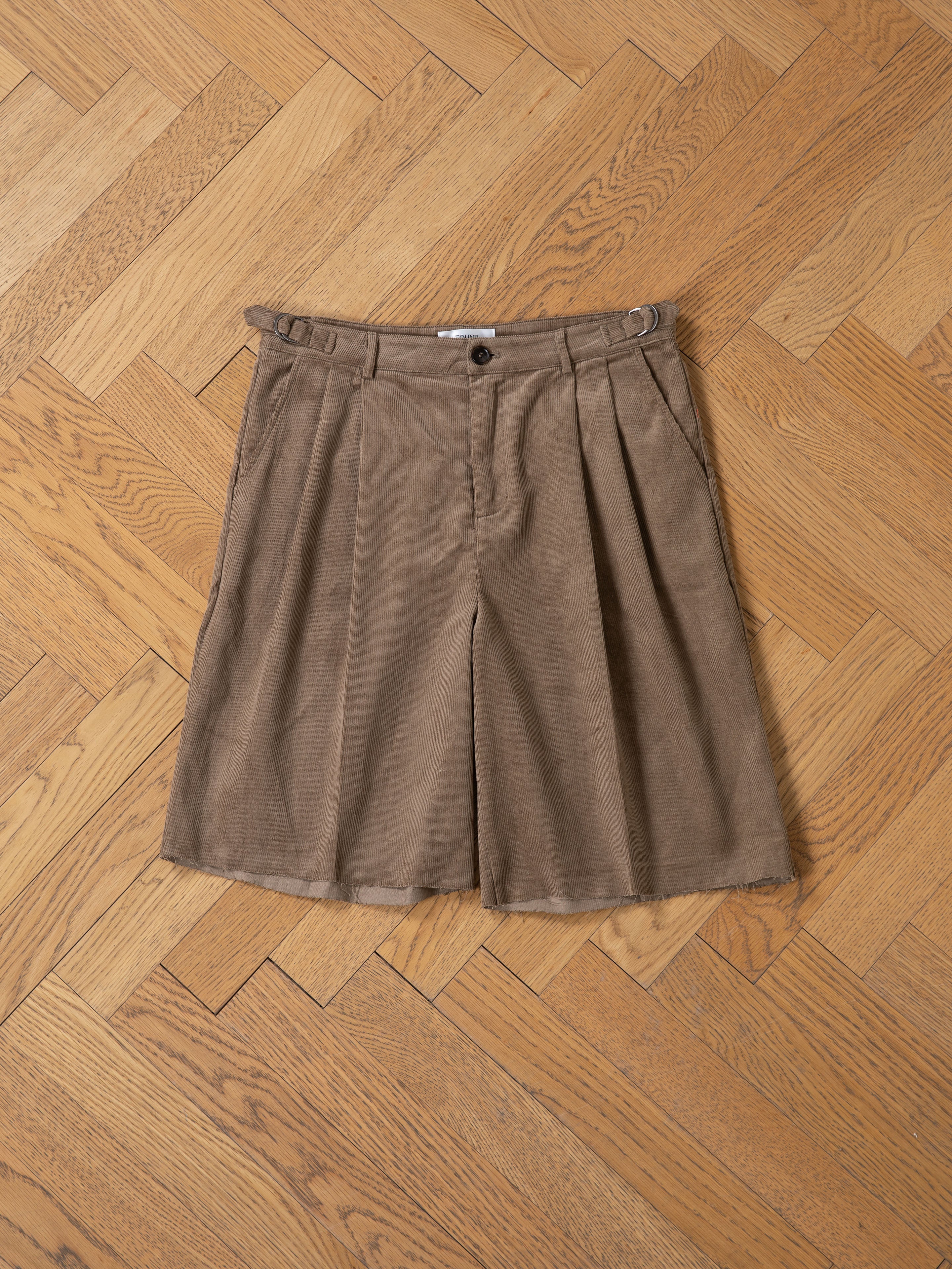 FOUND Corduroy Pleated Trouser Shorts feature a brown double-pleated design with belt loops, button, and zipper closure, shown on a wooden parquet floor.