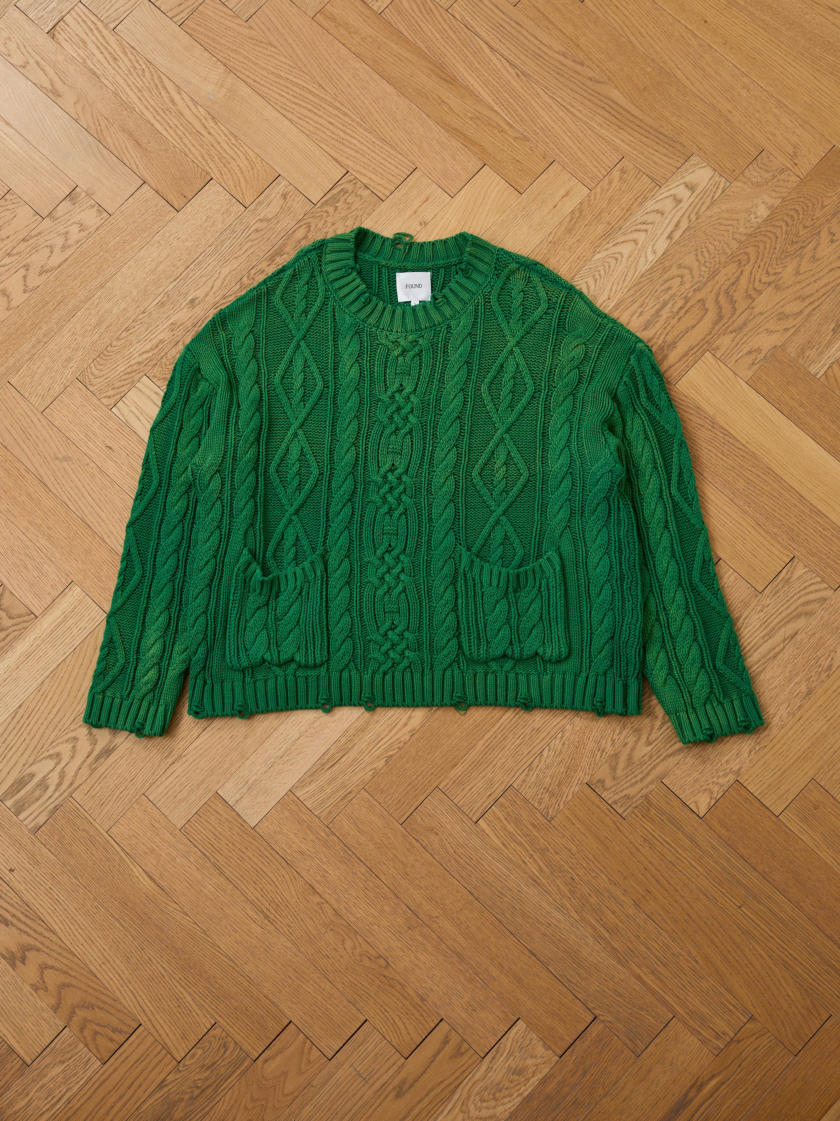 Laid flat on a wooden herringbone floor, the Found Cable Knit Pullover Sweater in green features two front pockets and a chunky knit design—this cozy unisex piece brings timeless style to any wardrobe.