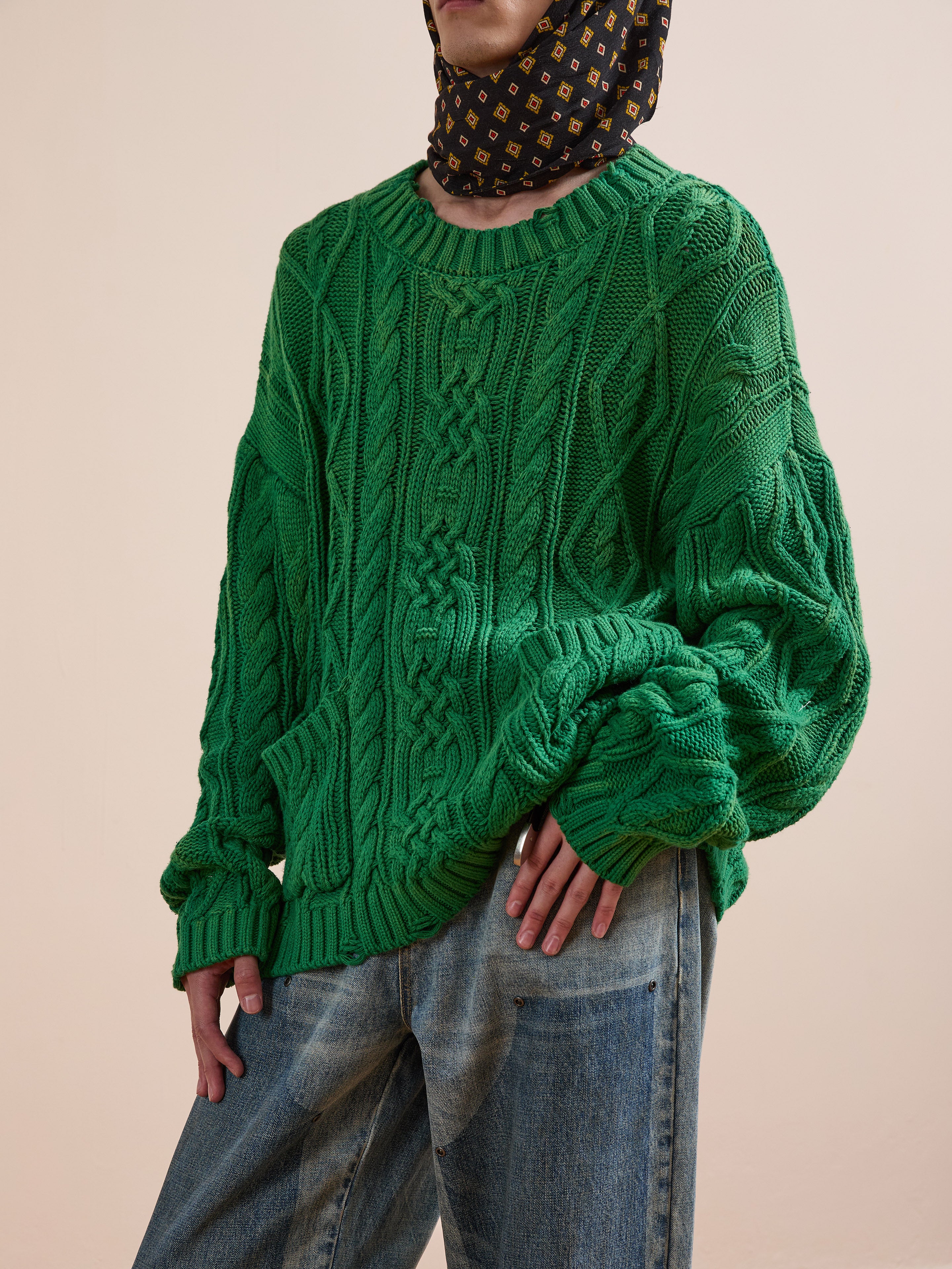 A person in the Found Cable Knit Pullover Sweater, styled with blue jeans and a patterned scarf, stands before a plain beige background.