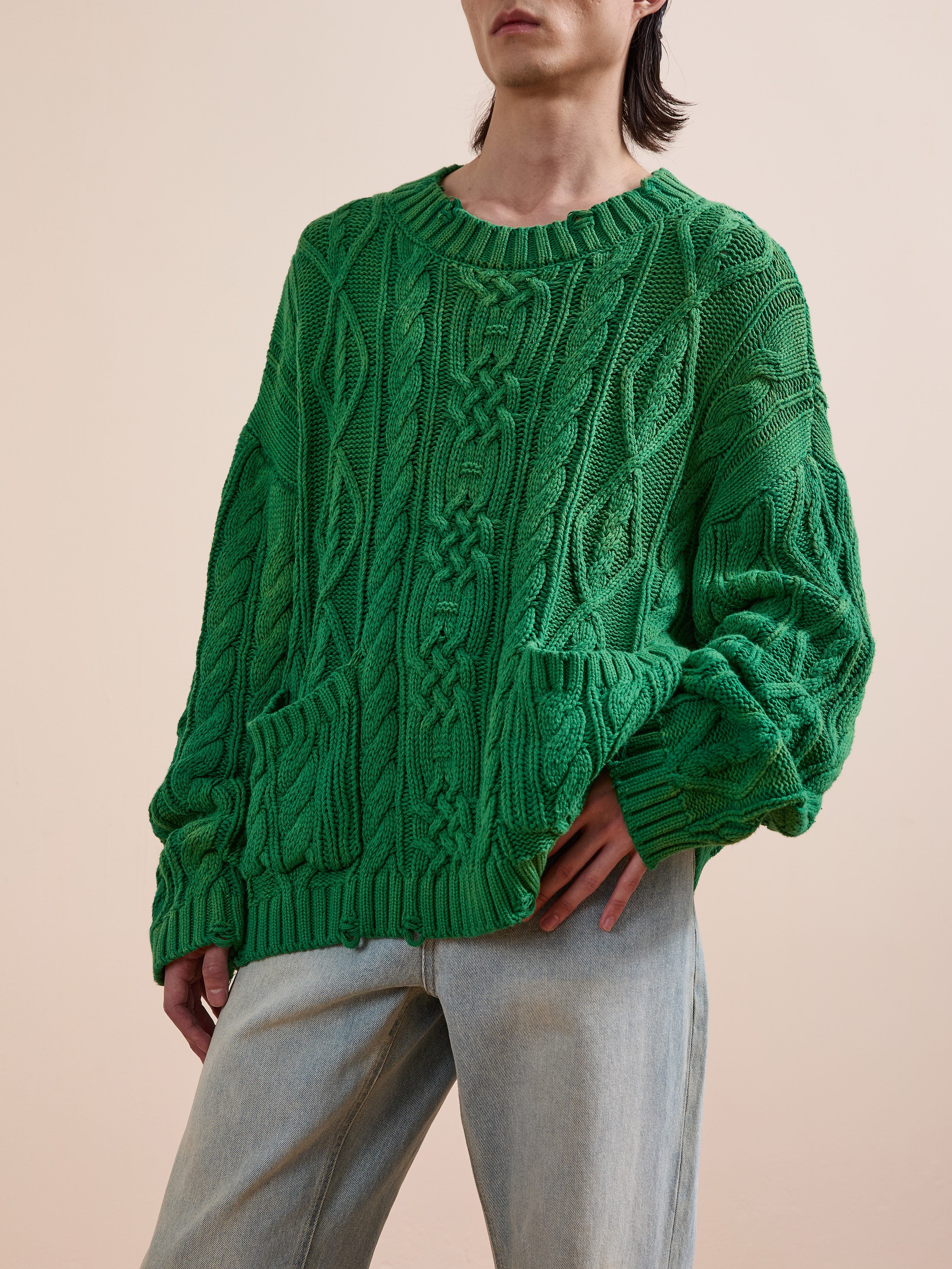 A person wears the Found Cable Knit Pullover Sweater in green with two front pockets, paired with light blue jeans, standing against a plain beige background.