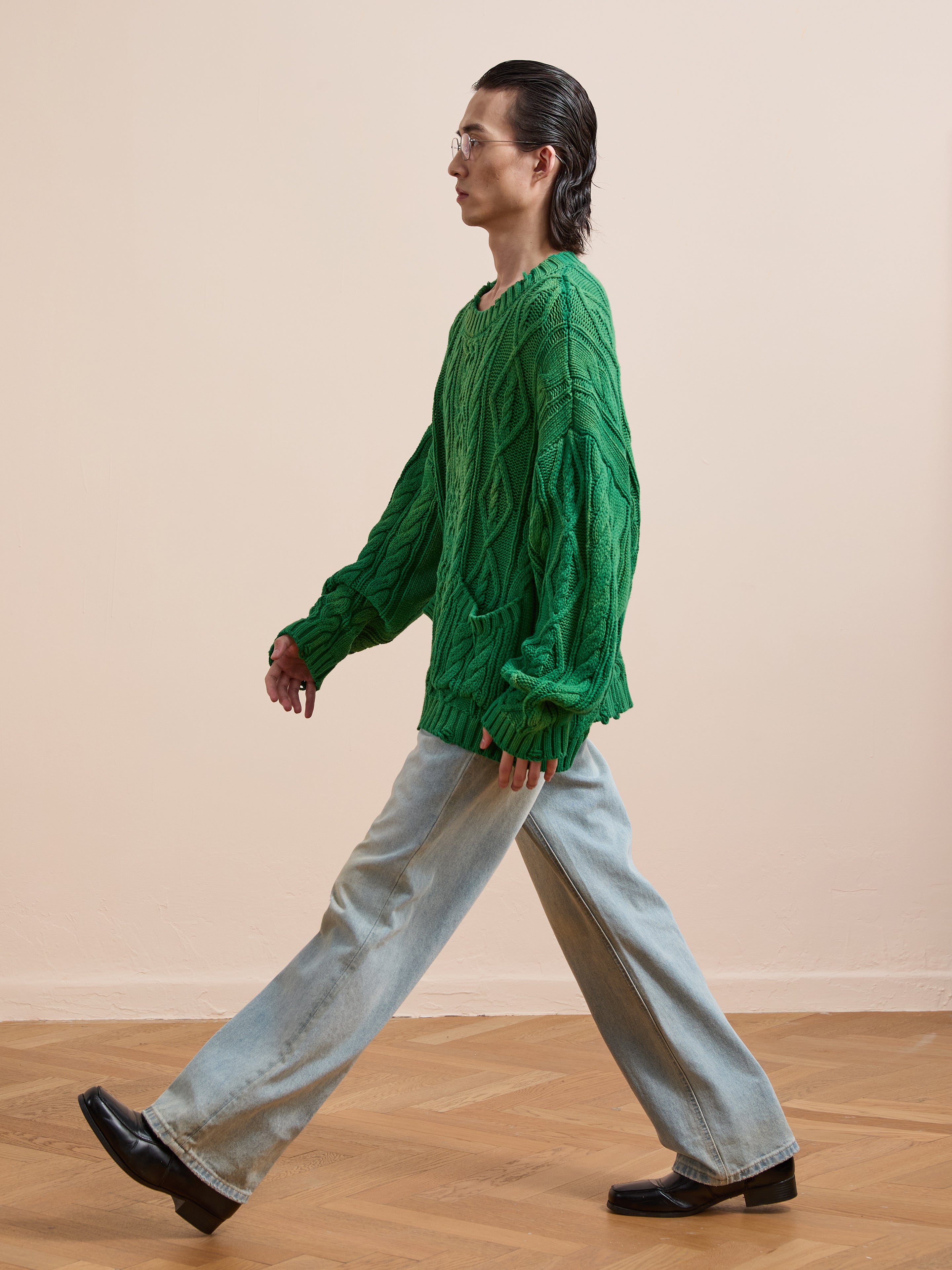 A person walks indoors on a wooden floor wearing the Found Cable Knit Pullover Sweater in green, paired with light blue wide-leg jeans and black shoes.