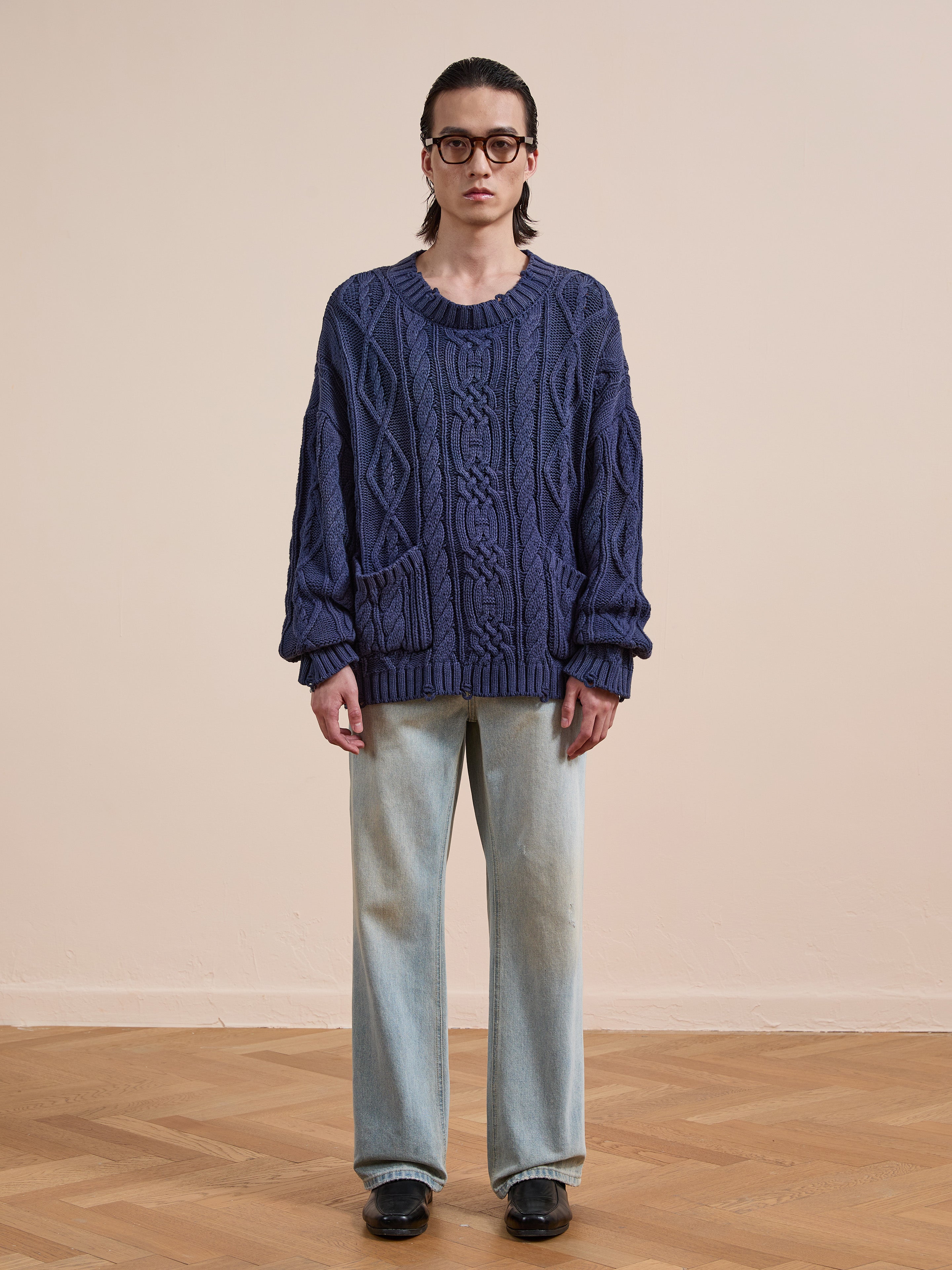 An individual wearing glasses, the Found Cable Knit Pullover Sweater in blue with pockets, paired with light-wash jeans and black shoes, stands indoors on a wooden floor against a plain background.