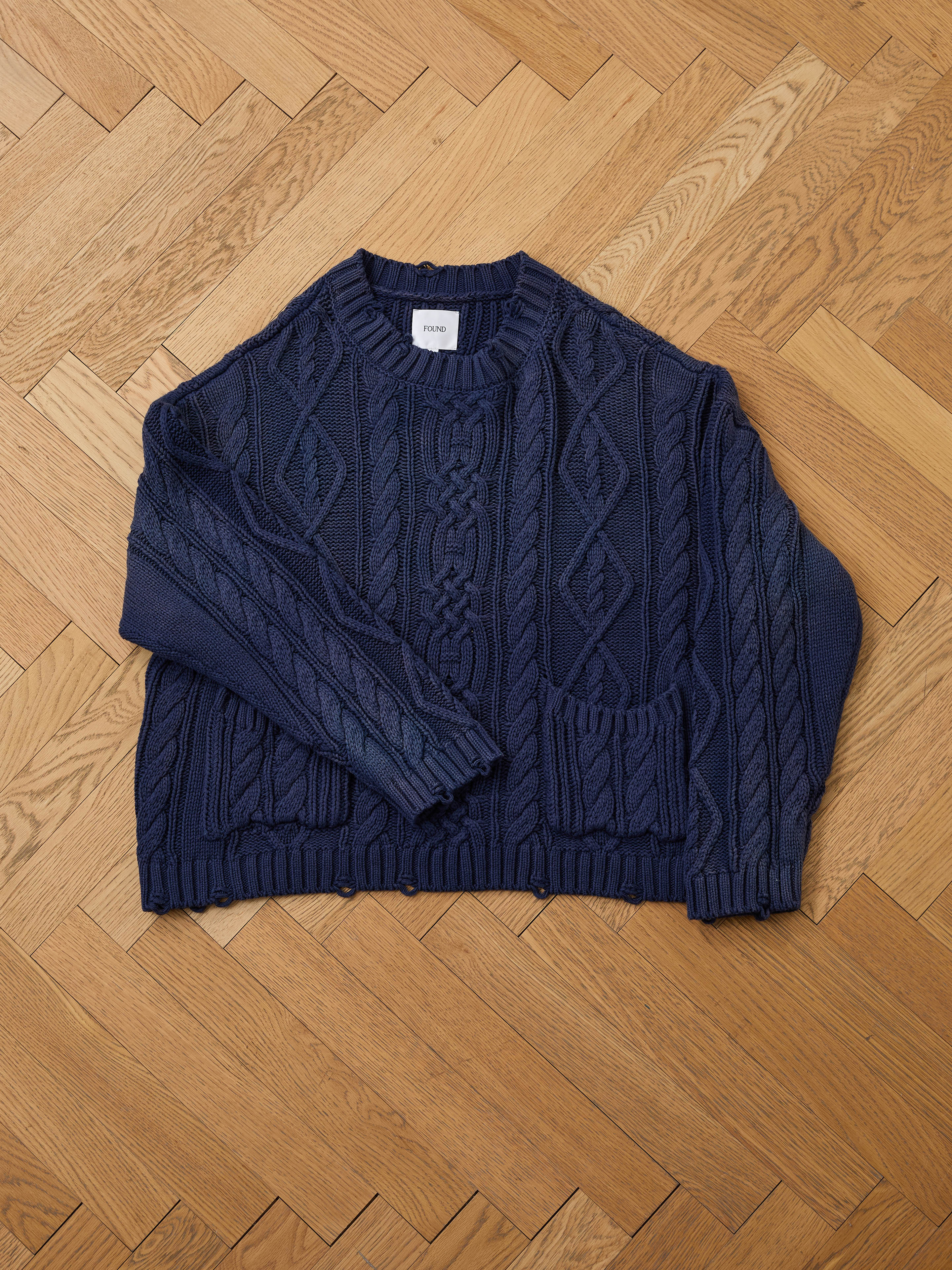 The Found Cable Knit Pullover Sweater, a unisex navy design with long sleeves and two front pockets, is laid flat on a herringbone wooden floor.