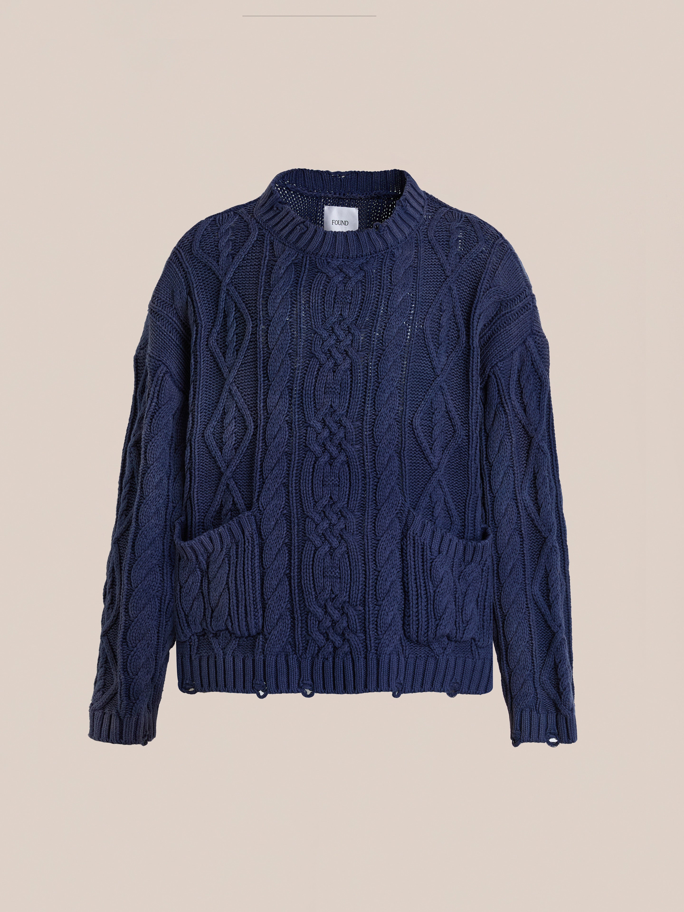 The Found Cable Knit Pullover Sweater features a navy blue design with long sleeves, round neckline, and two front pockets—an essential unisex style—shown against a plain beige background.
