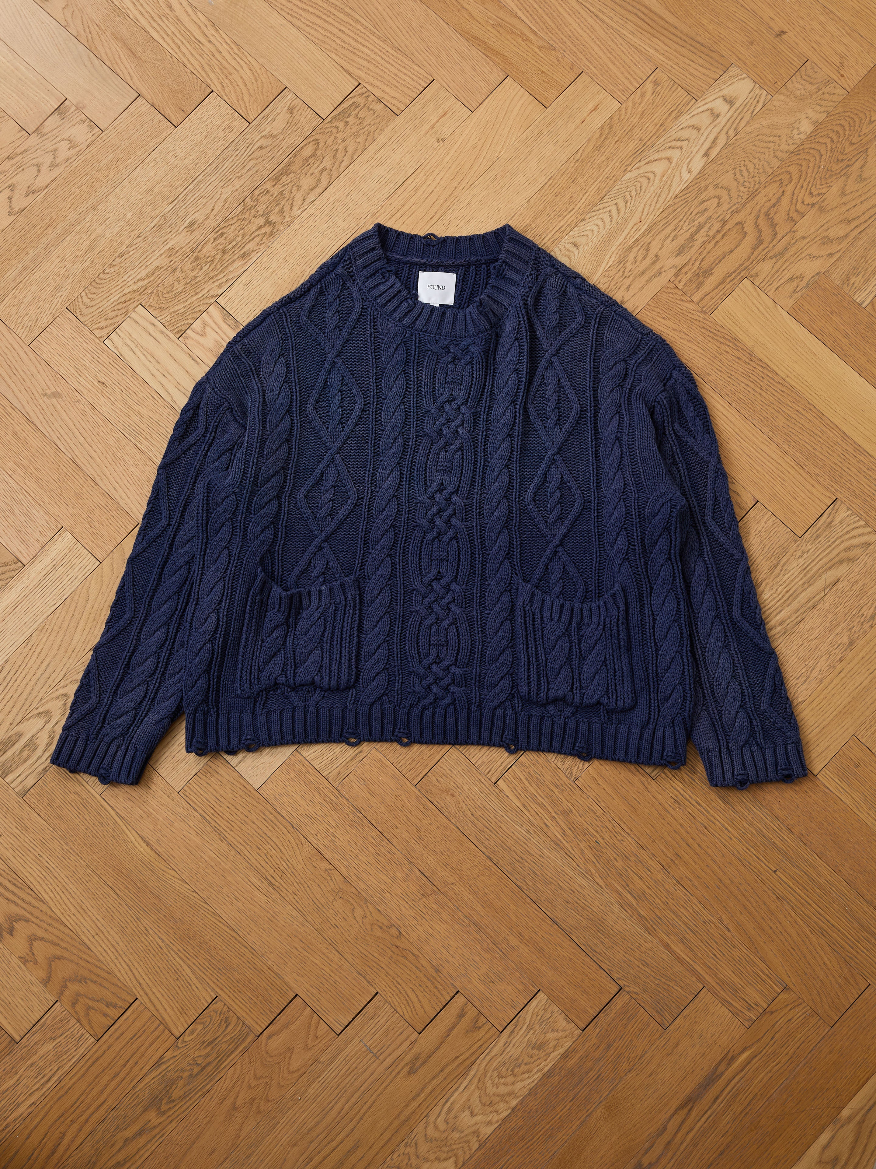 The Found Cable Knit Pullover Sweater, a navy blue unisex style with two front pockets, is shown laid flat on a light brown herringbone wood floor.