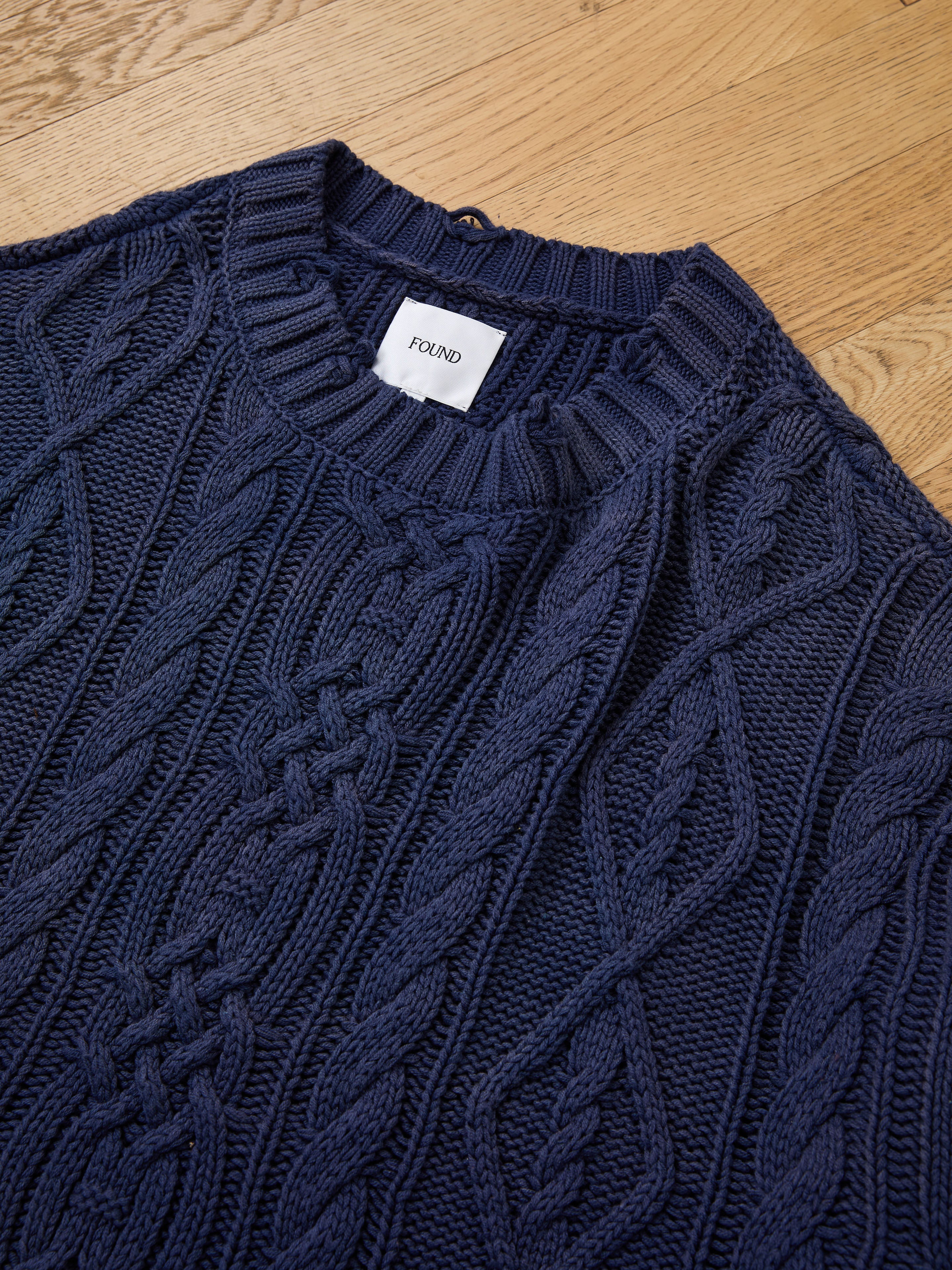 The Found Cable Knit Pullover Sweater in navy blue is laid flat on a wooden surface. This unisex sweater features classic cable knit styling and timeless comfort.
