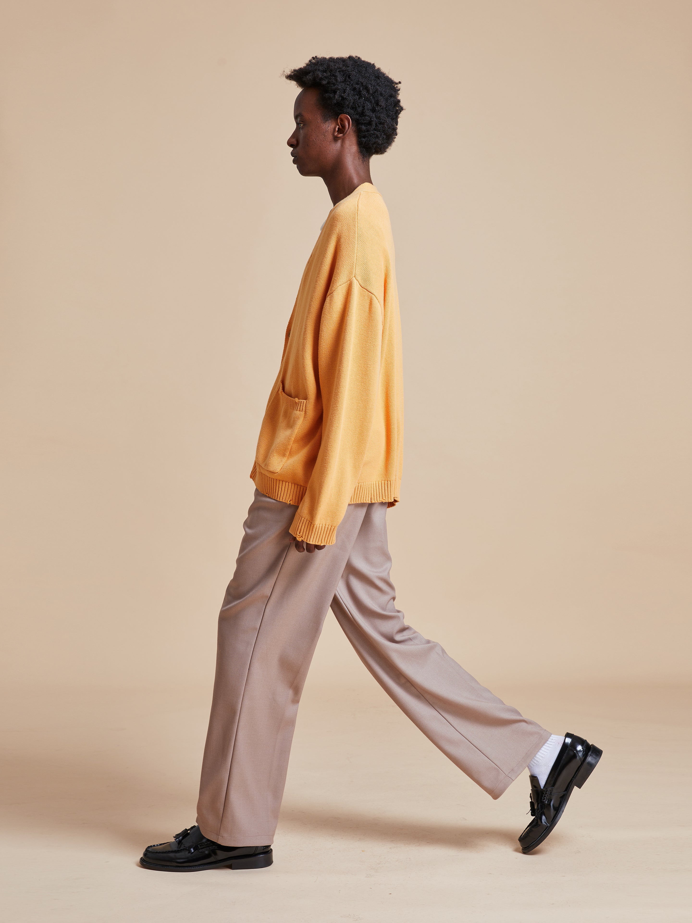 A man in a Found Cadmium Distressed Cardigan and trousers walking on a beige background.