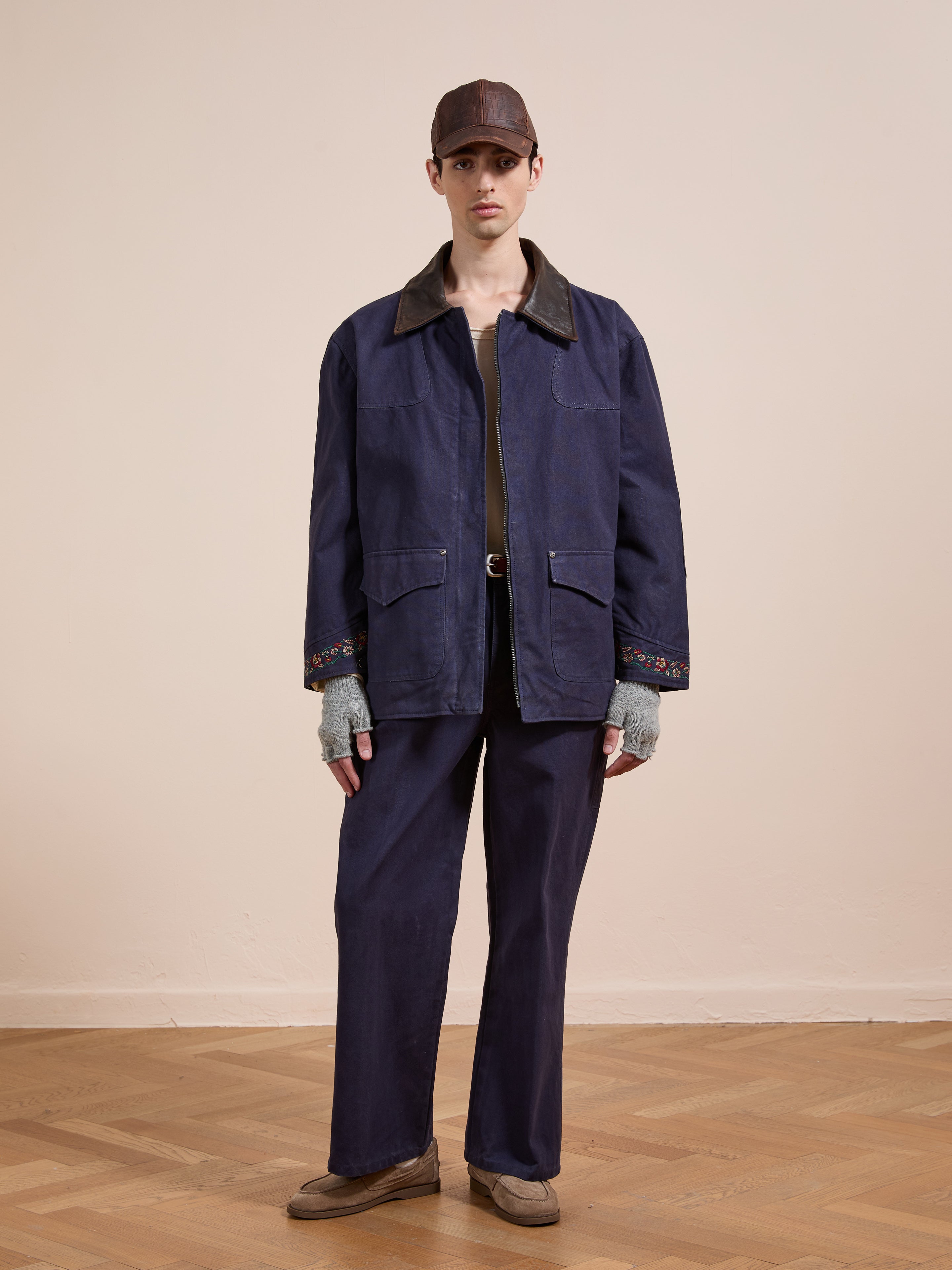 A person wears Found's Canvas Distressed Barn Jacket with a leather collar, wide-leg navy pants, brown shoes, gray fingerless gloves, and a brown cap indoors on a wood floor. Unisex relaxed fit offers timeless style.