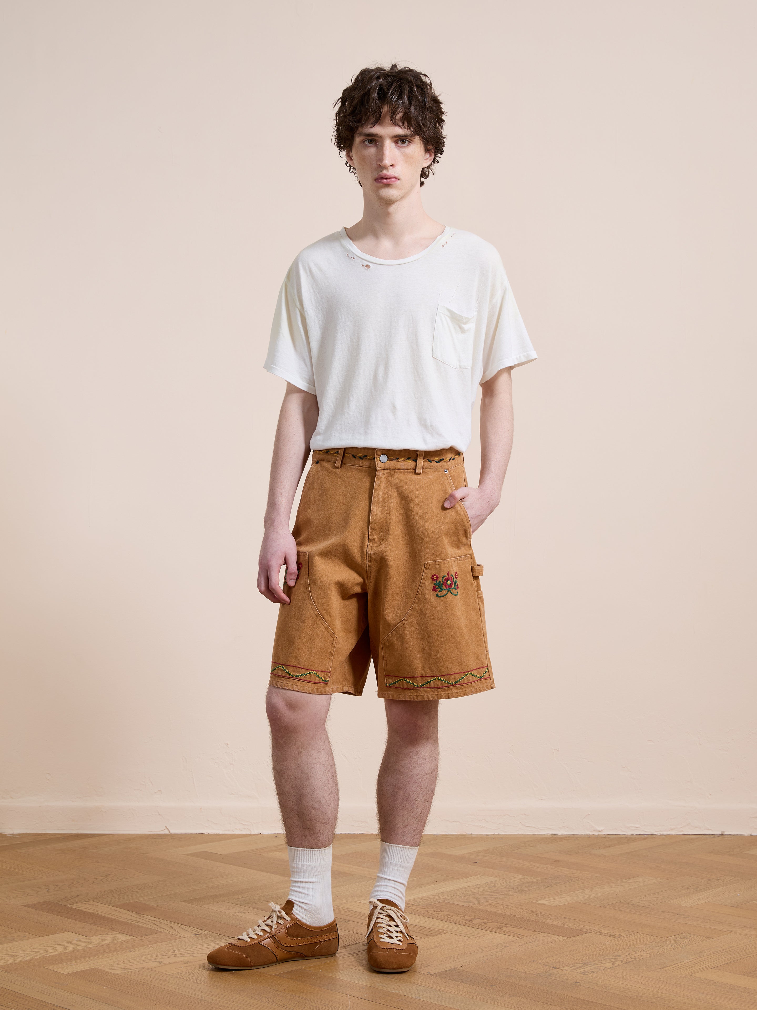 Indoors on a wooden floor, a person wears a loose white t-shirt, Found Canvas Embroidered Shorts in tan knee-length style, white socks, and brown sneakers.