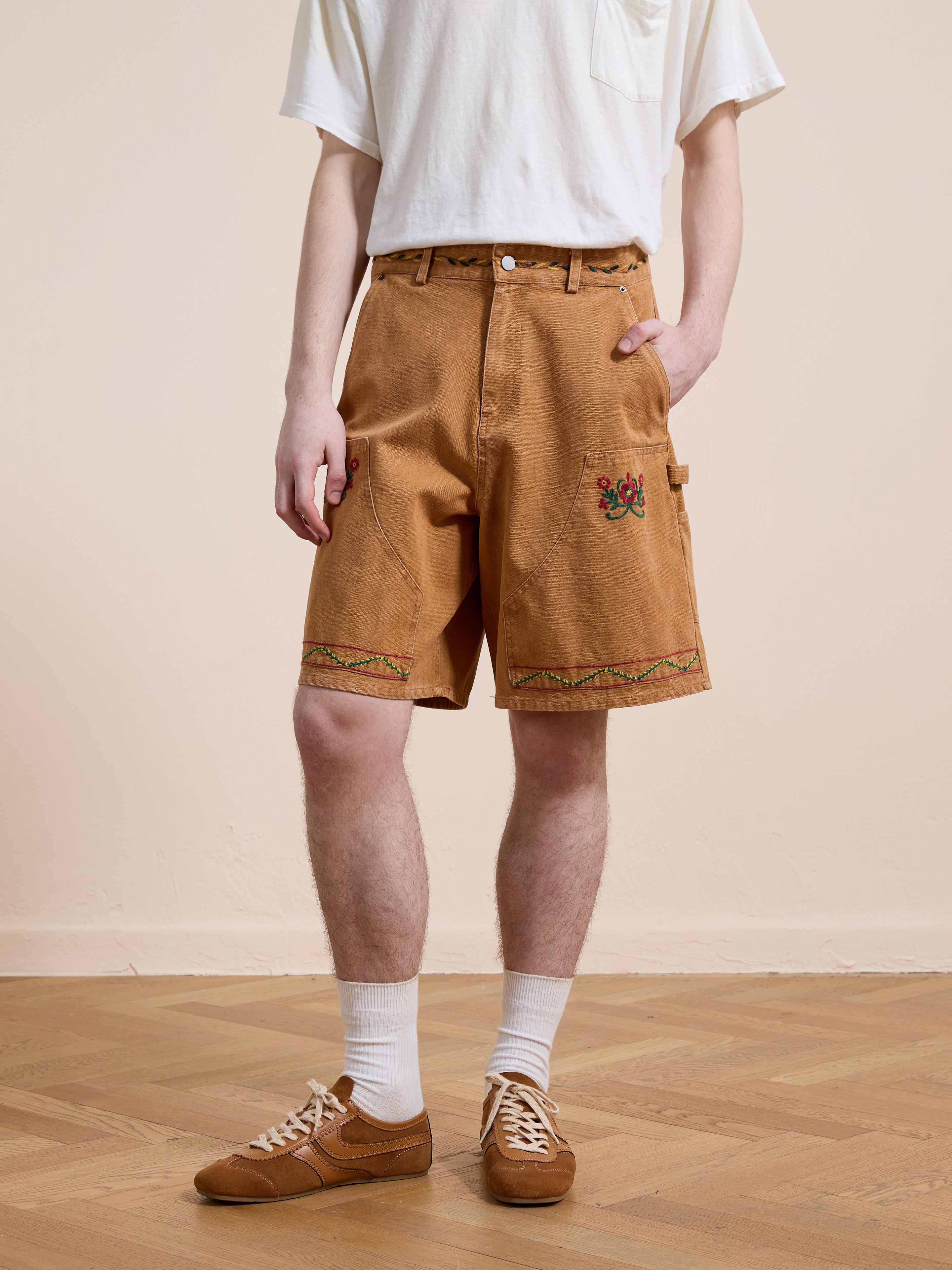 A person indoors wears a white tee, Found Canvas Embroidered Shorts, white socks, and brown sneakers on a wooden floor—a modern twist on unisex workwear.