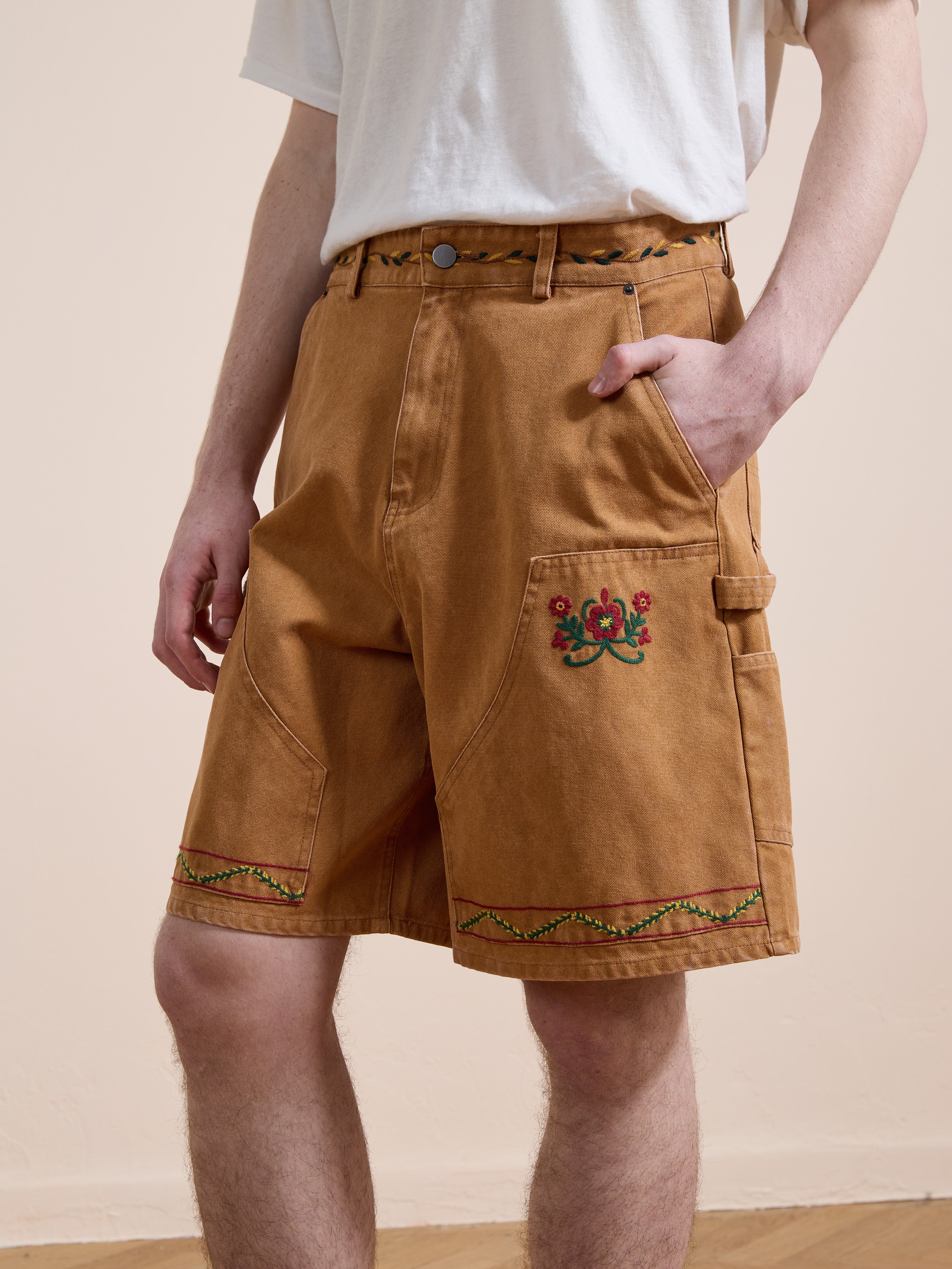 A person wears Found Canvas Embroidered Shorts with a white T-shirt, standing with one hand in their pocket.