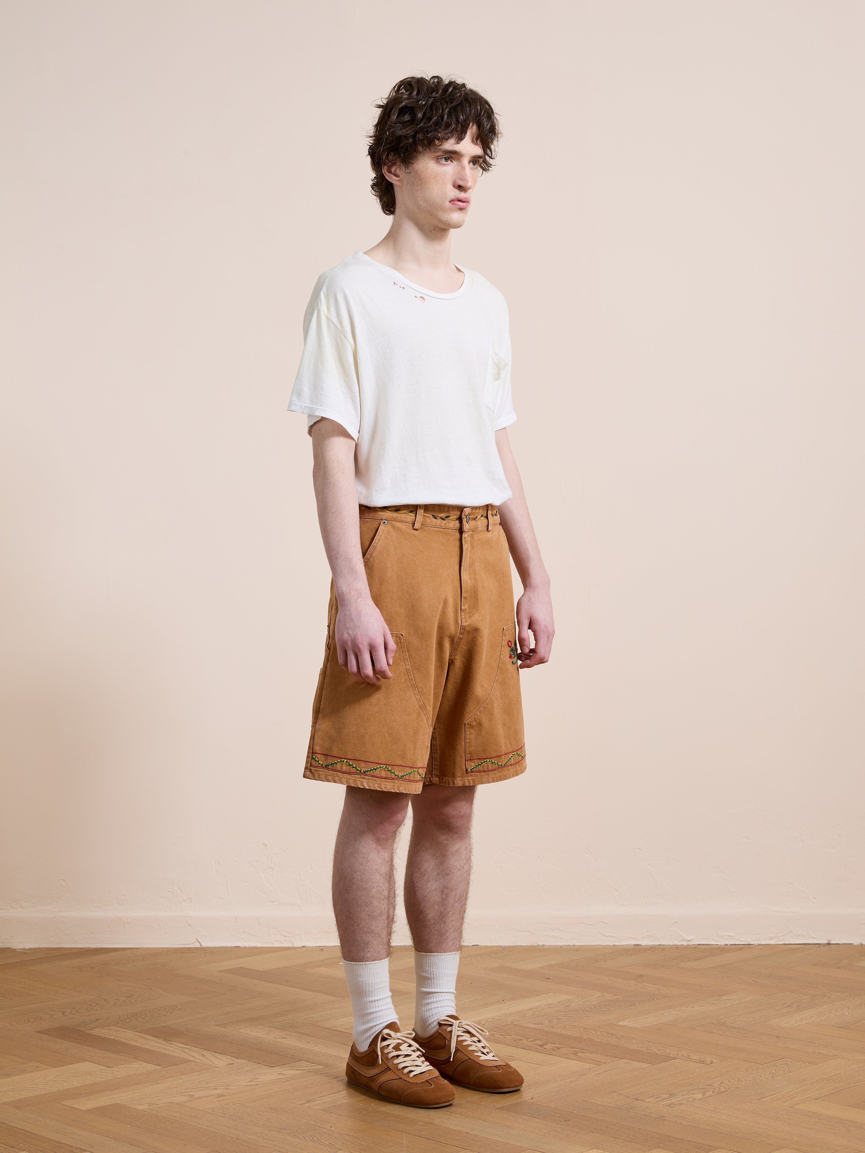 A person stands indoors on a parquet floor against a beige wall, wearing a white t-shirt, Found Canvas Embroidered Shorts, white socks, and brown shoes.