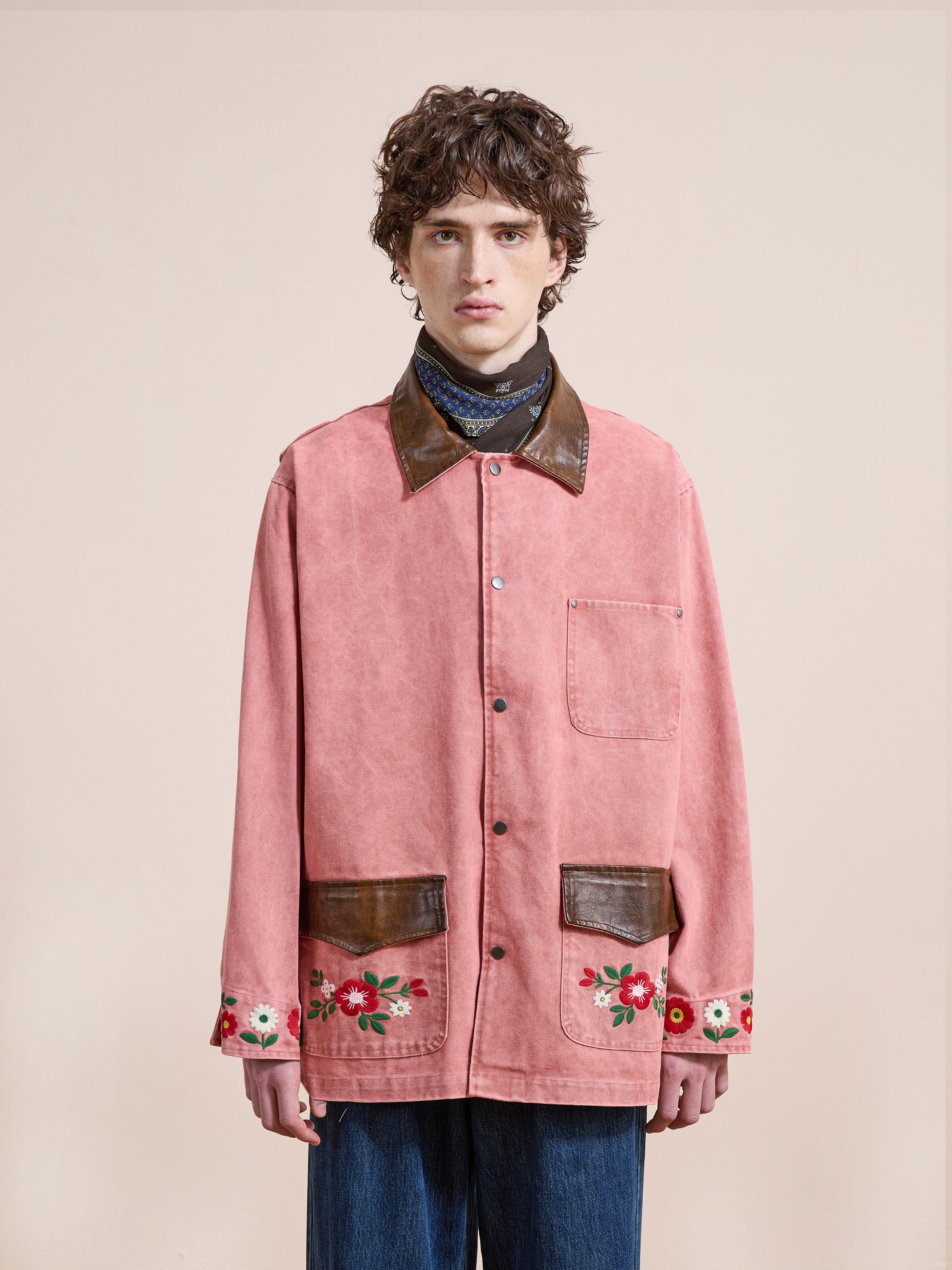 A person is wearing the Found Canvas Embroidered Barn Jacket, featuring a pink relaxed fit, brown leather collar and pockets, floral embroidery on the sleeves, paired with blue jeans against a plain background.
