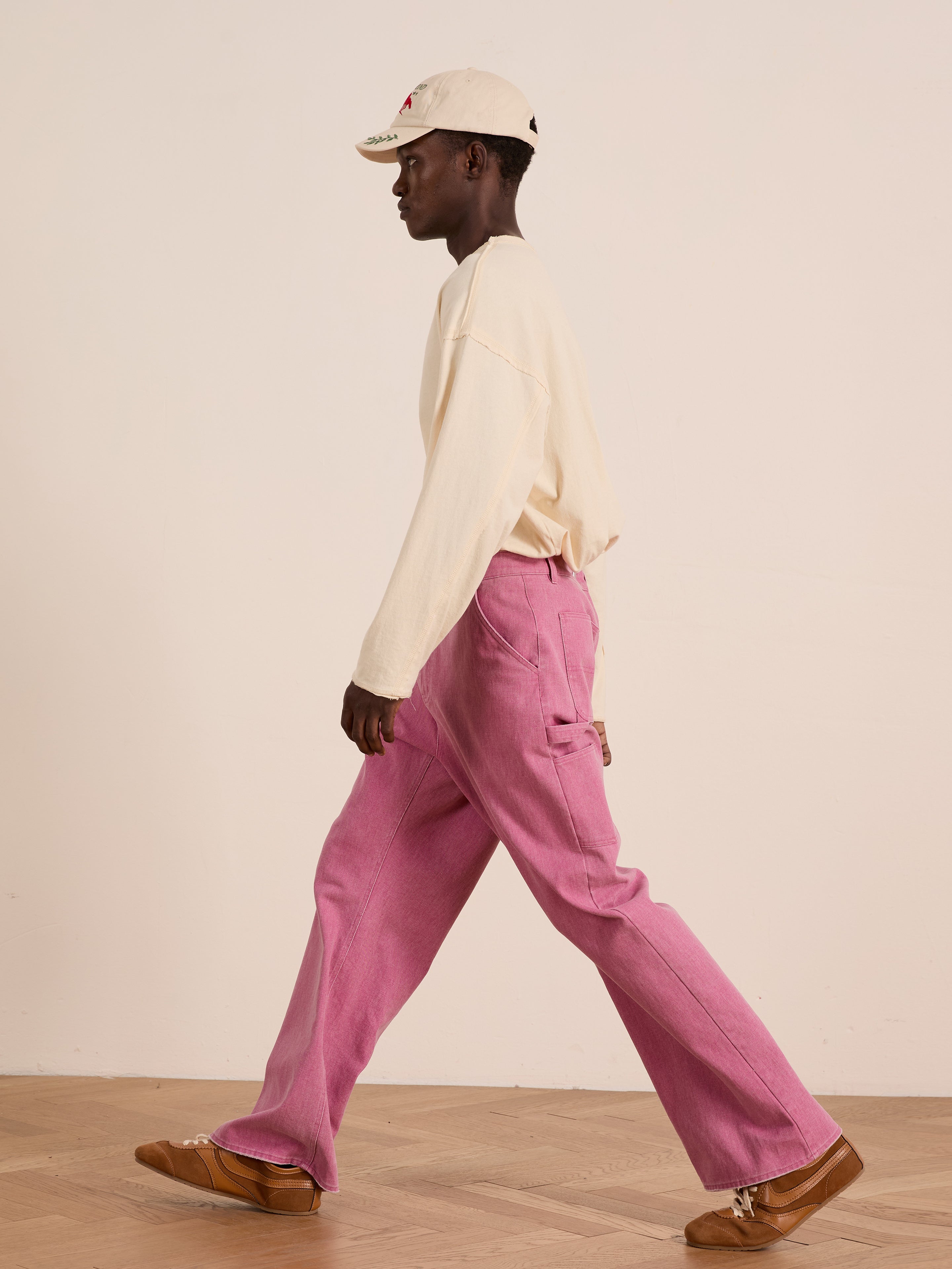 A person models FOUND Flare Work Pants in pink with a cream long-sleeve shirt, tan shoes, and a cream baseball cap, walking indoors on a wooden floor by a plain wall.