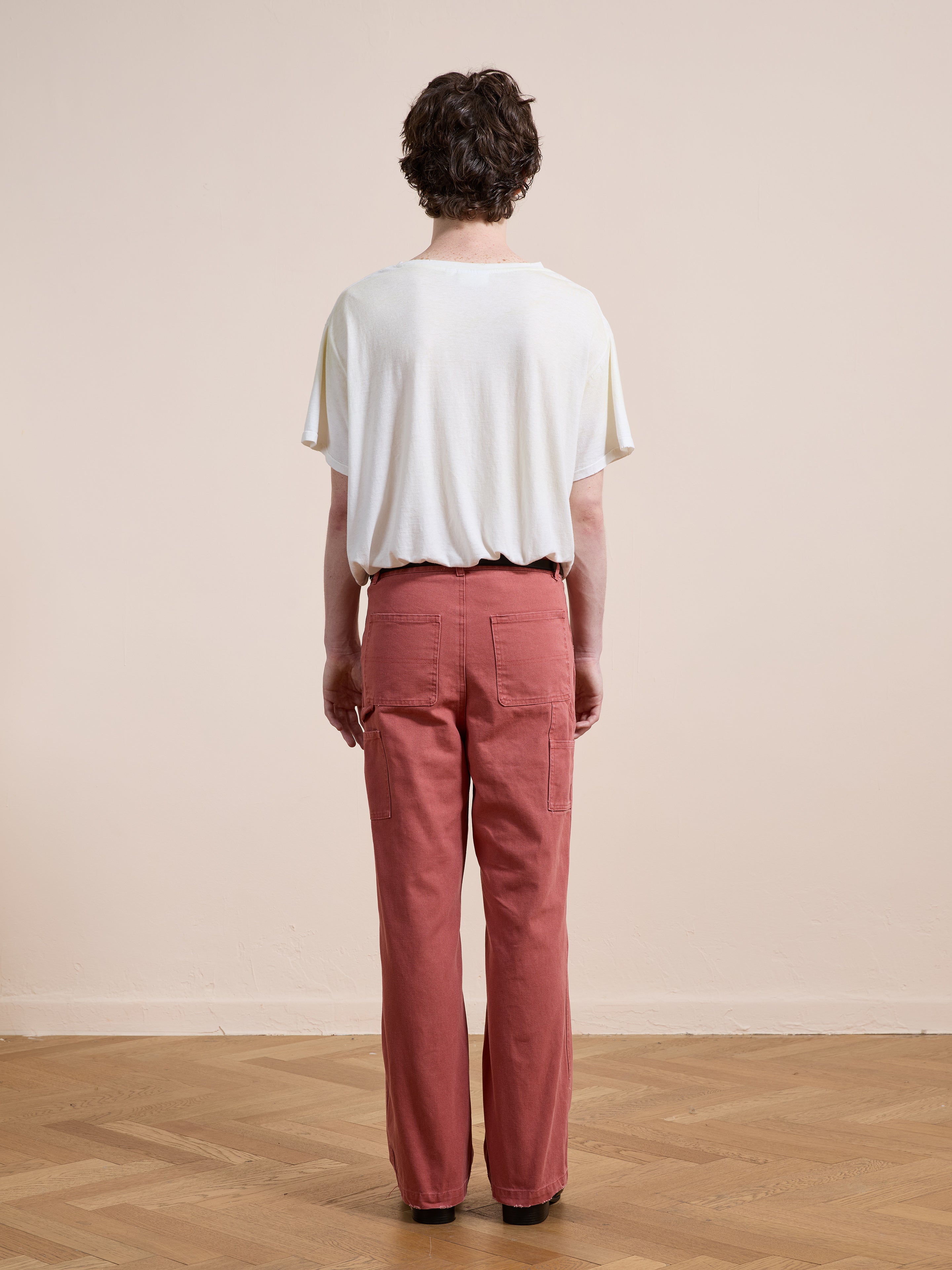 A person stands indoors on a wooden floor, facing away from the camera, wearing a loose white T-shirt and FOUND Canvas Faded Pants.
