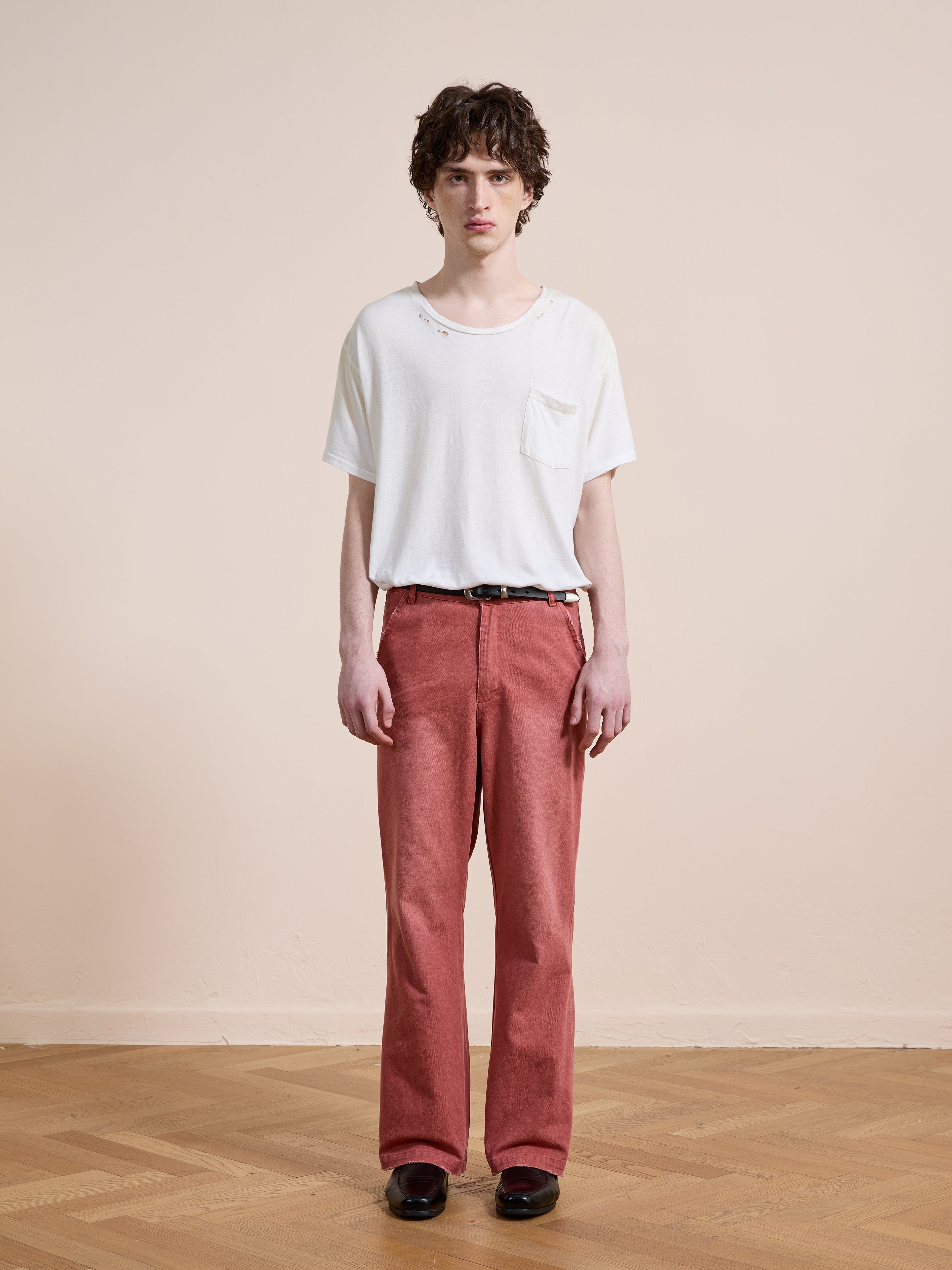A person stands indoors against a plain beige wall, wearing a white t-shirt, FOUND Canvas Faded Pants in vintage salmon wash, and black shoes on a light wooden floor.