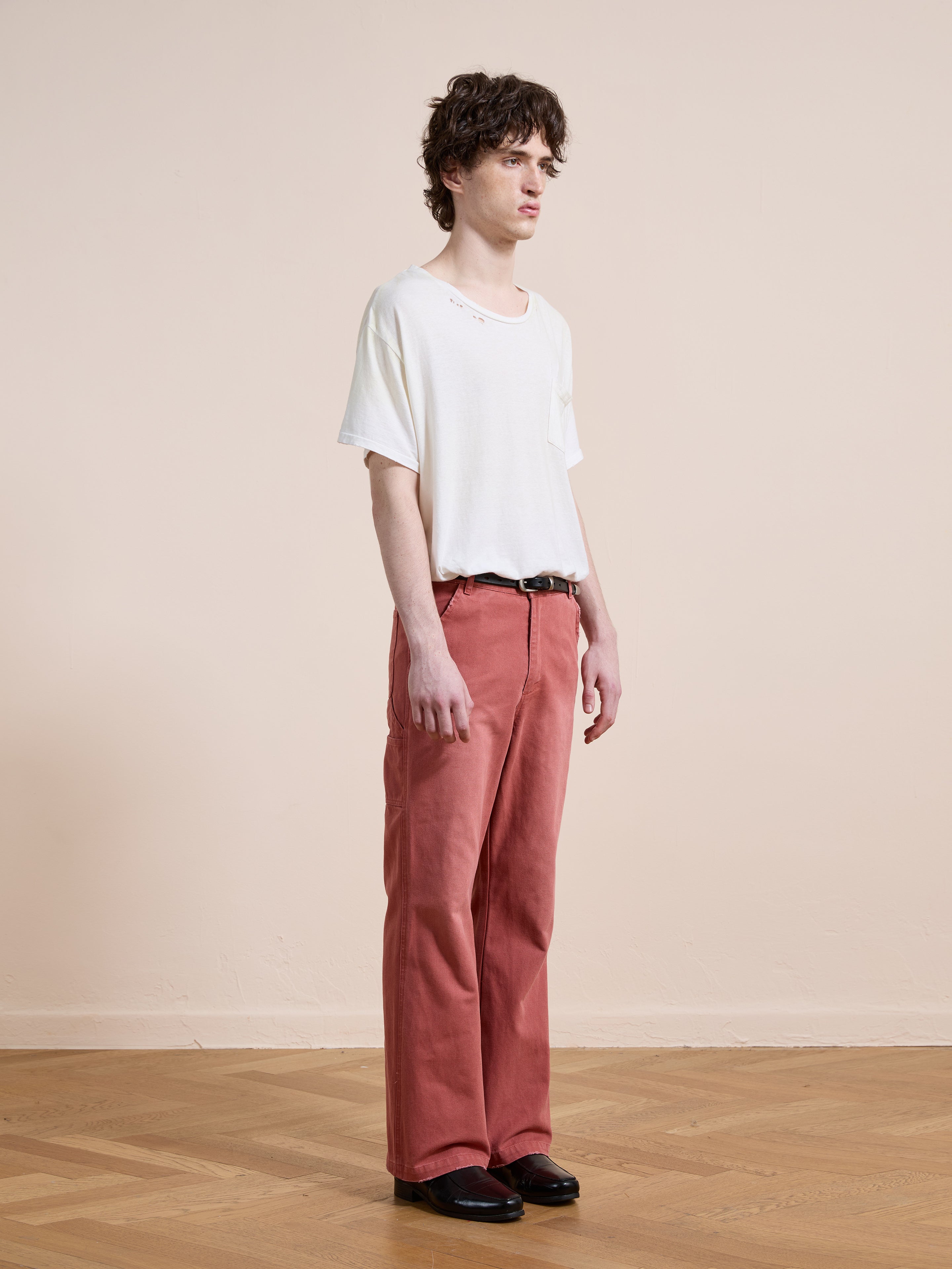 A person stands indoors on a wooden floor against a beige wall, wearing a plain white t-shirt, FOUND Canvas Faded Pants in red, a black belt, and black shoes.