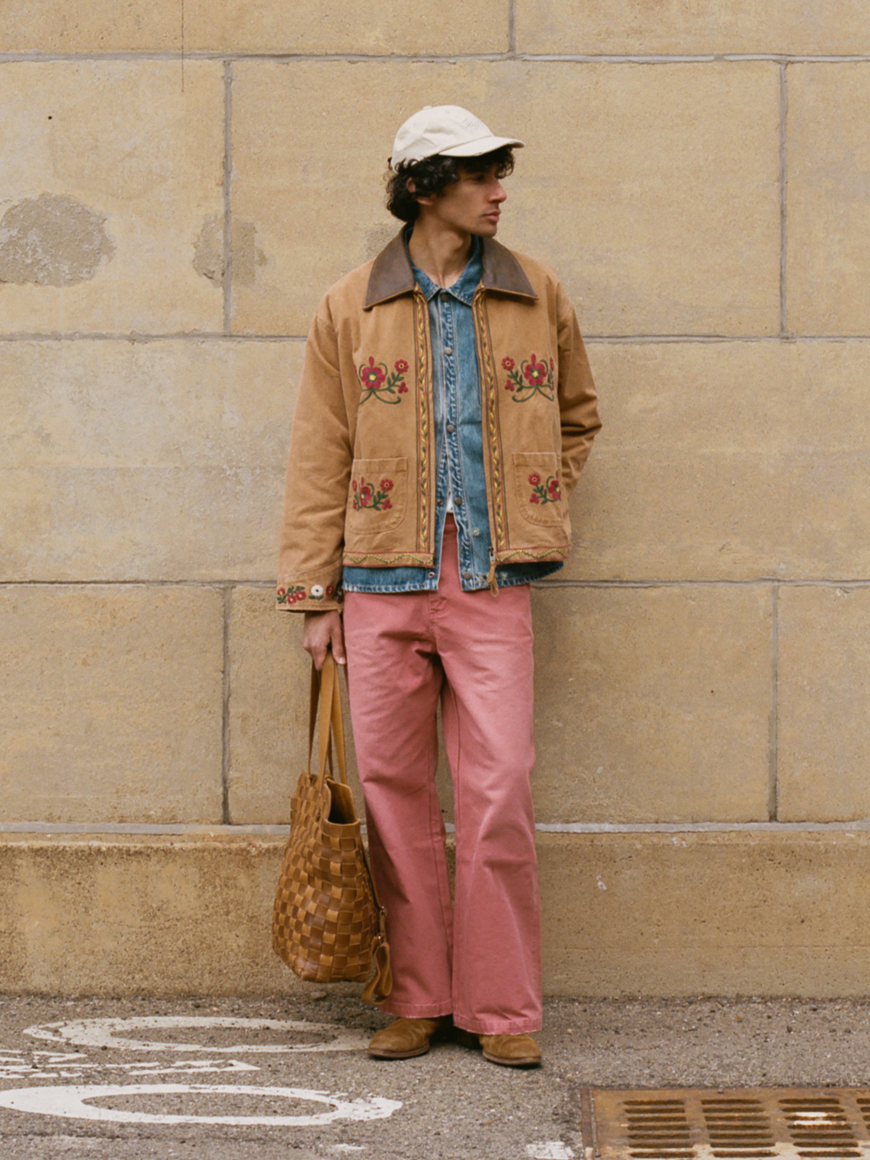 A person stands against a stone wall wearing the Found Rosette Canvas Work Jacket over a denim shirt, pink pants, brown shoes, a white cap, and holding a woven tote bag.