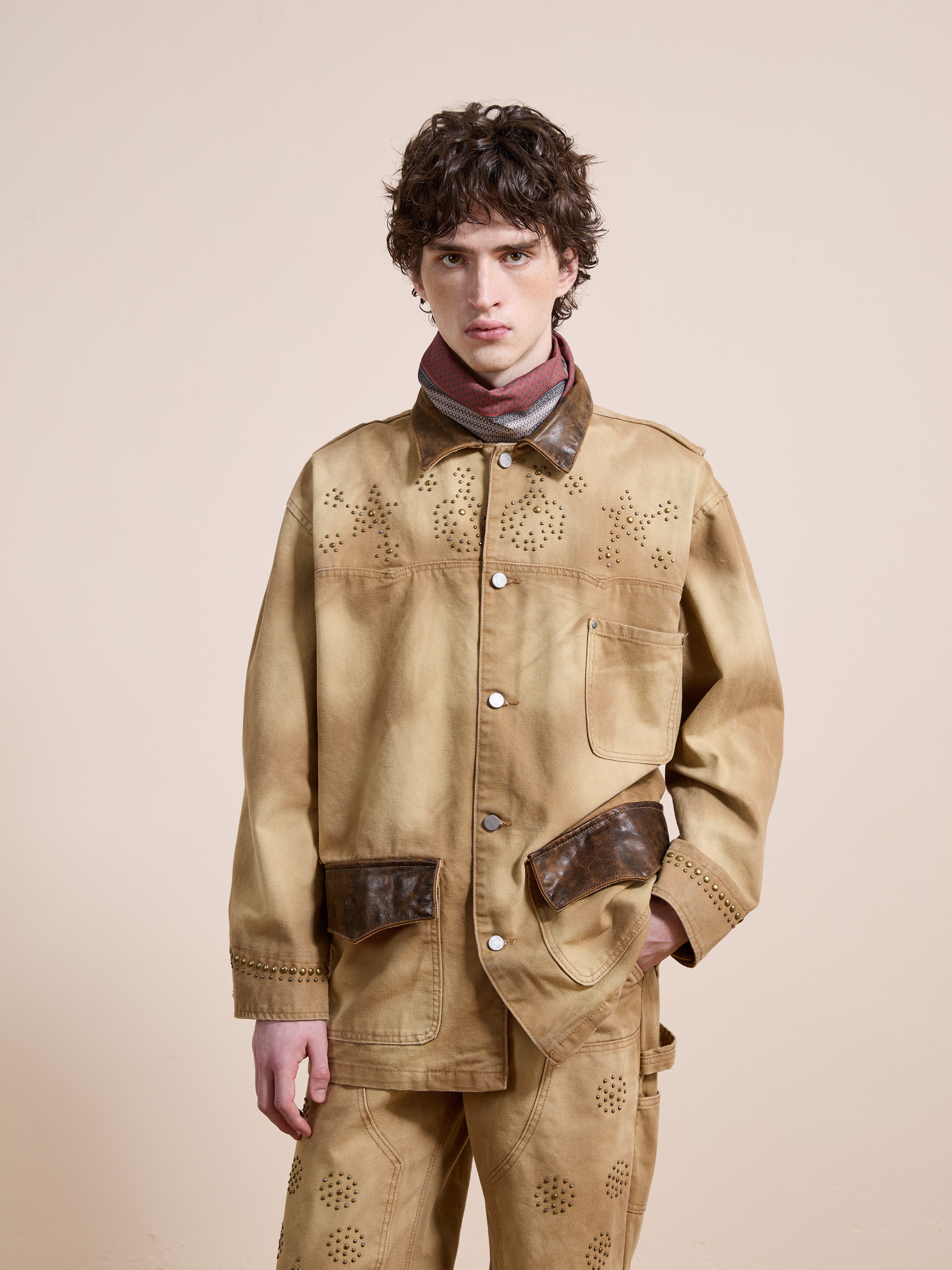 A person with short curly hair wears the Found Canvas Studded Work Jacket in beige with brown accents and matching pants, standing against a plain light background.