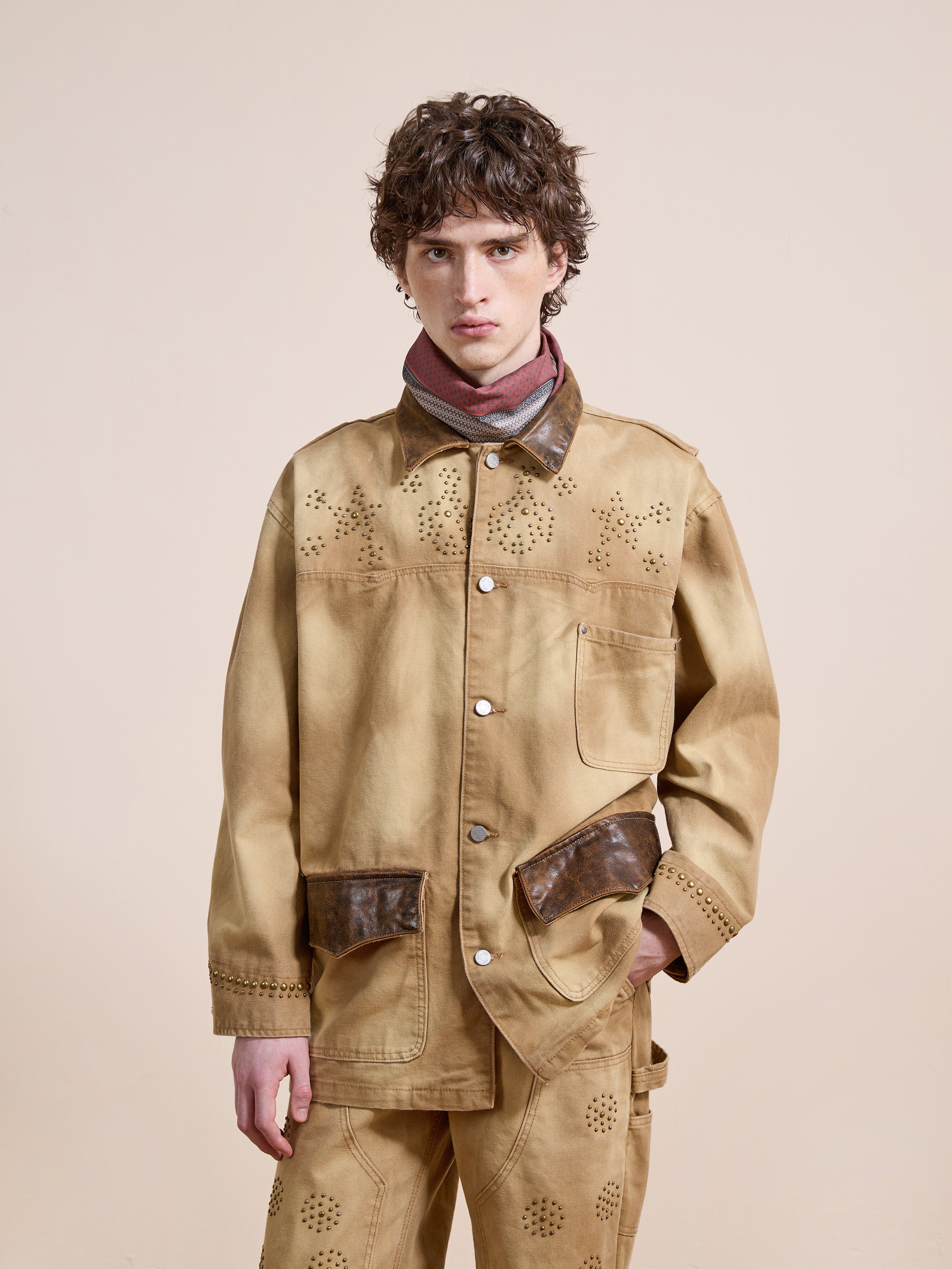 A person wears the Found Canvas Studded Barn Jacket with brown leather details, matching pants, and a pink and gray scarf, standing against a plain beige background.