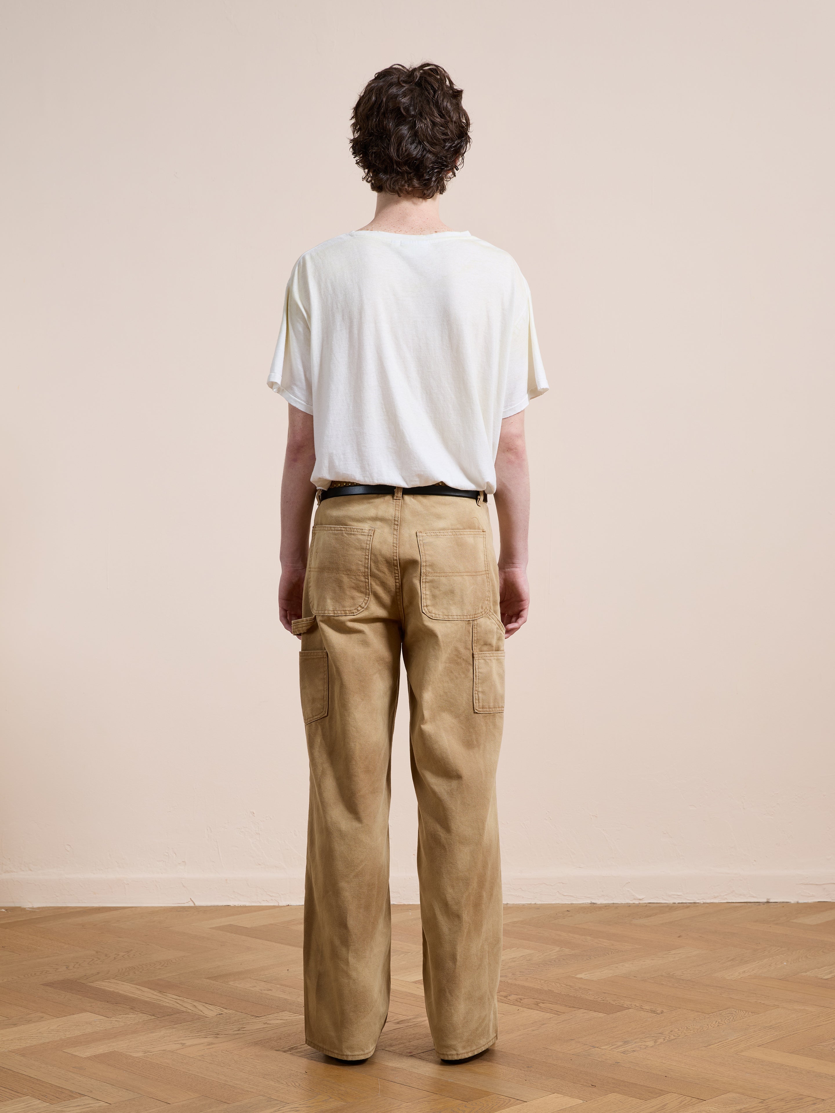 A person stands indoors on a wooden floor, facing away from the camera and wearing FOUND Canvas Studded Work Pants with a white t-shirt, highlighting classic unisex workwear style.