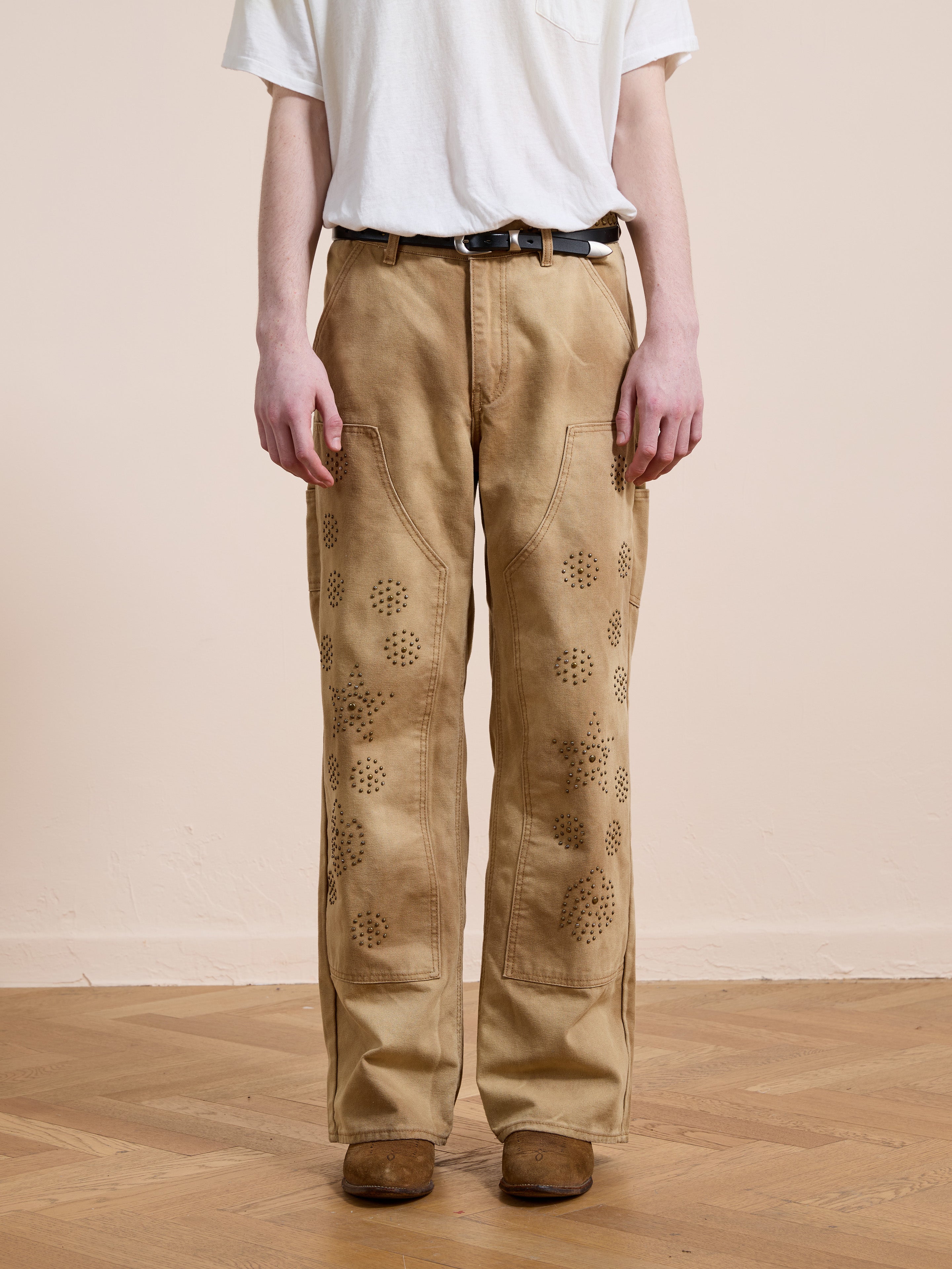 A person stands on a wooden floor against a plain light-colored wall, wearing FOUND Canvas Studded Work Pants with a patterned design, a white t-shirt, and brown shoes.