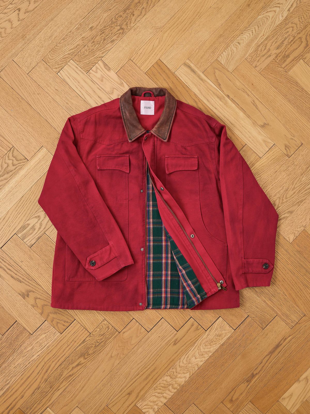 A Found Canvas Utility Barn Jacket in red, featuring a brown corduroy collar, is laid flat on a wooden floor and partially unbuttoned to display its green and red plaid lining.