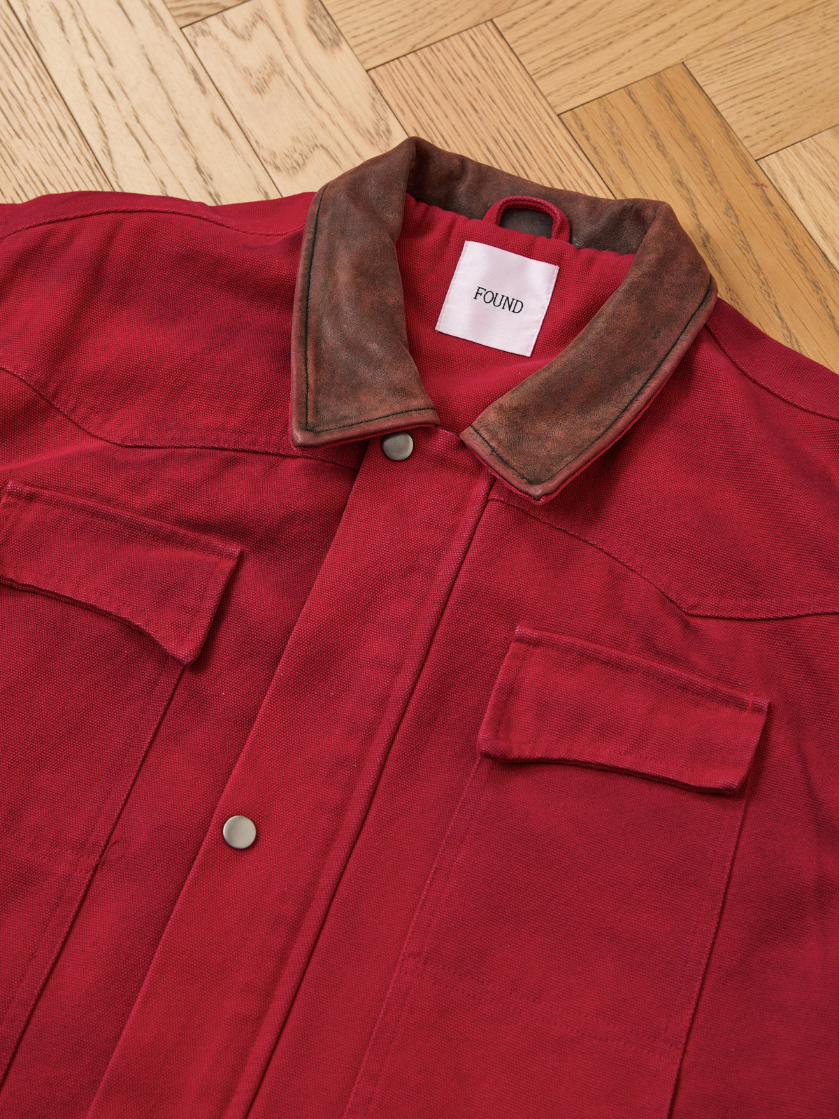 The Canvas Utility Barn Jacket by Found, featuring a red canvas construction with two chest pockets and a brown corduroy collar, is laid flat on a wooden floor.