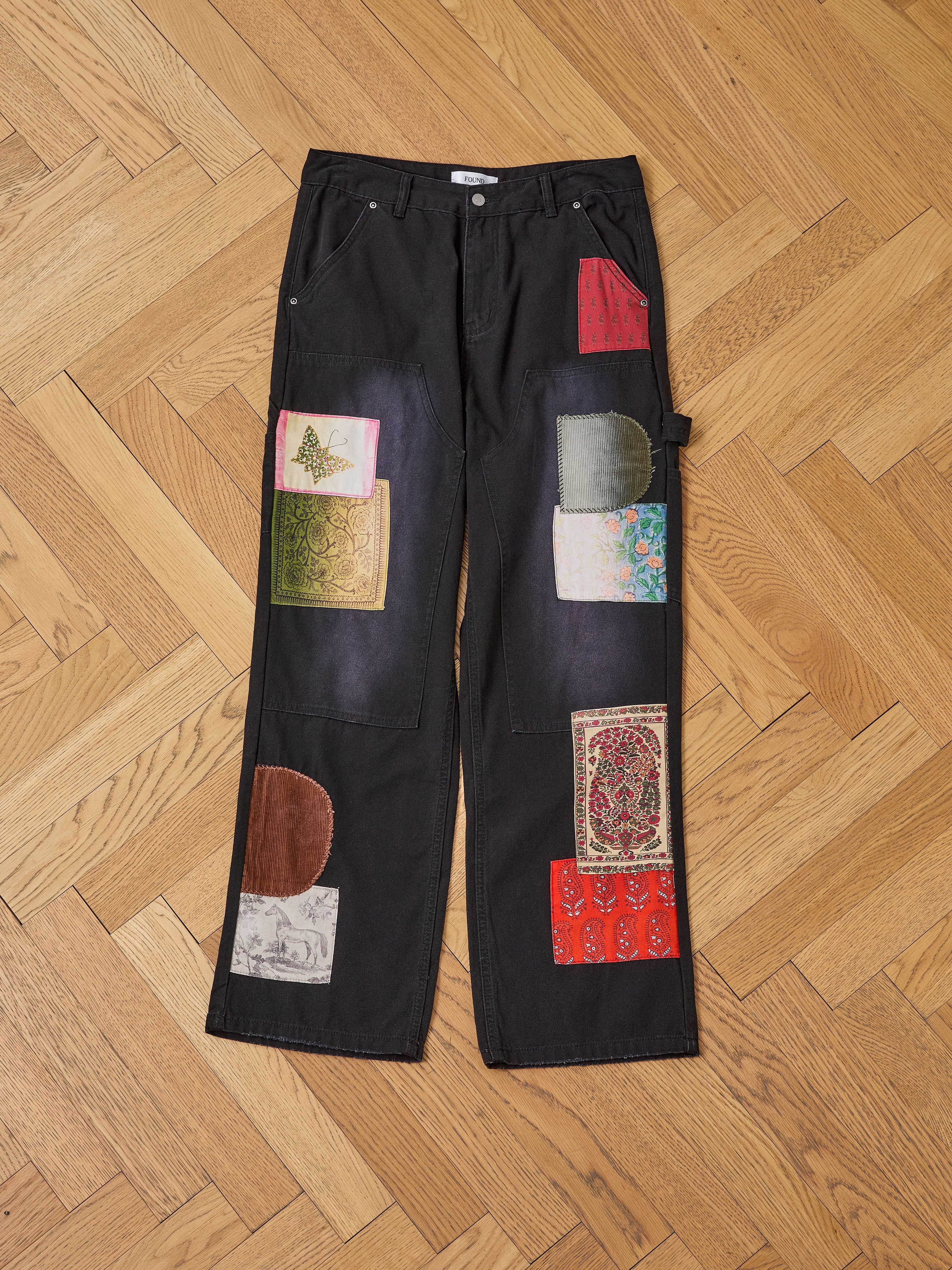 FOUND’s Tapestry Carpenter Canvas Pants feature colorful fabric patches inspired by Pakistani quilts, sewn onto the legs and displayed flat on a wooden herringbone floor.
