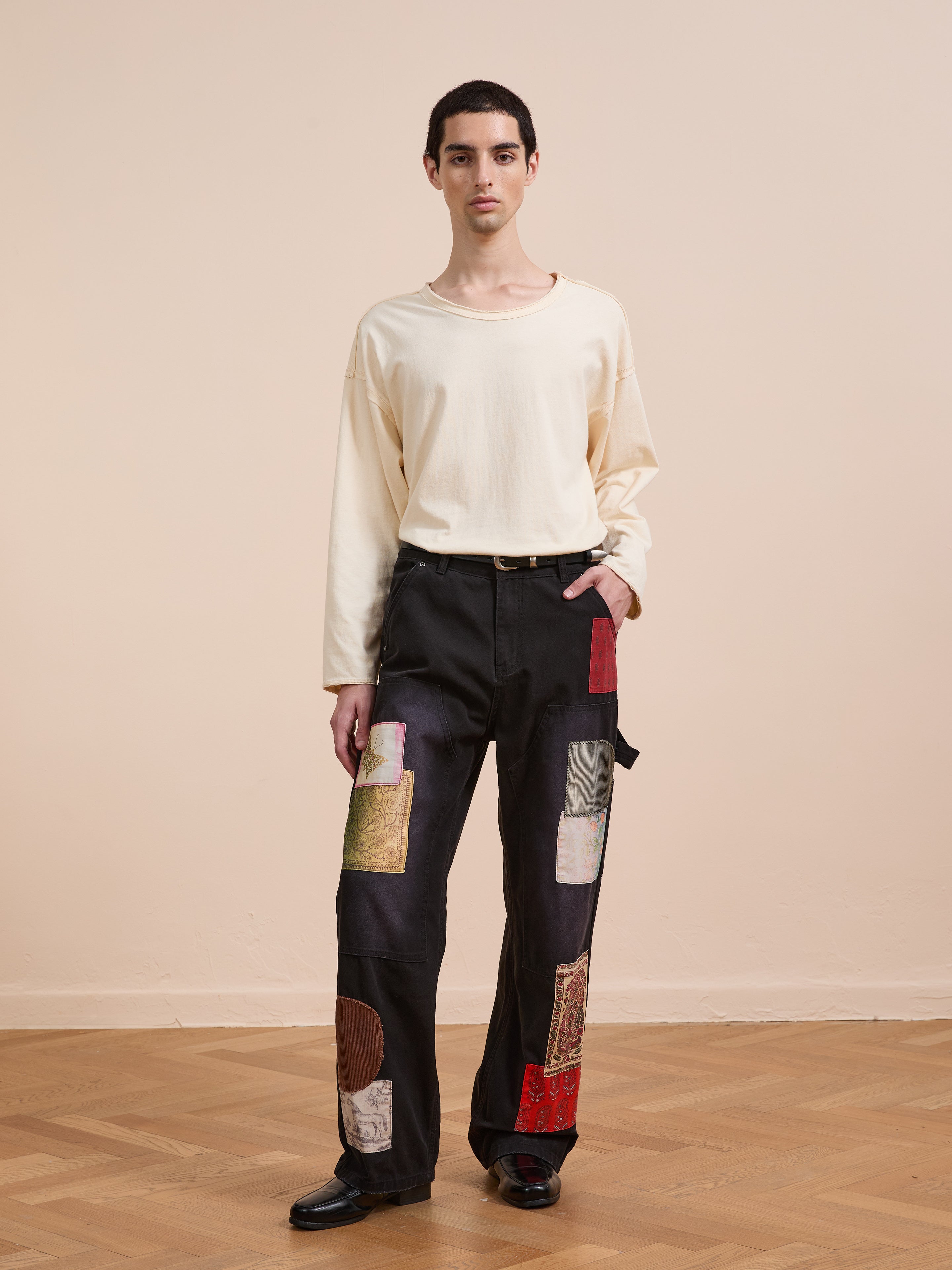 A person stands indoors wearing a loose cream long-sleeve shirt and FOUND Tapestry Carpenter Canvas Pants—unisex, relaxed fit black pants featuring bold, colorful Pakistani quilt-inspired patchwork details, with one hand in their pocket.