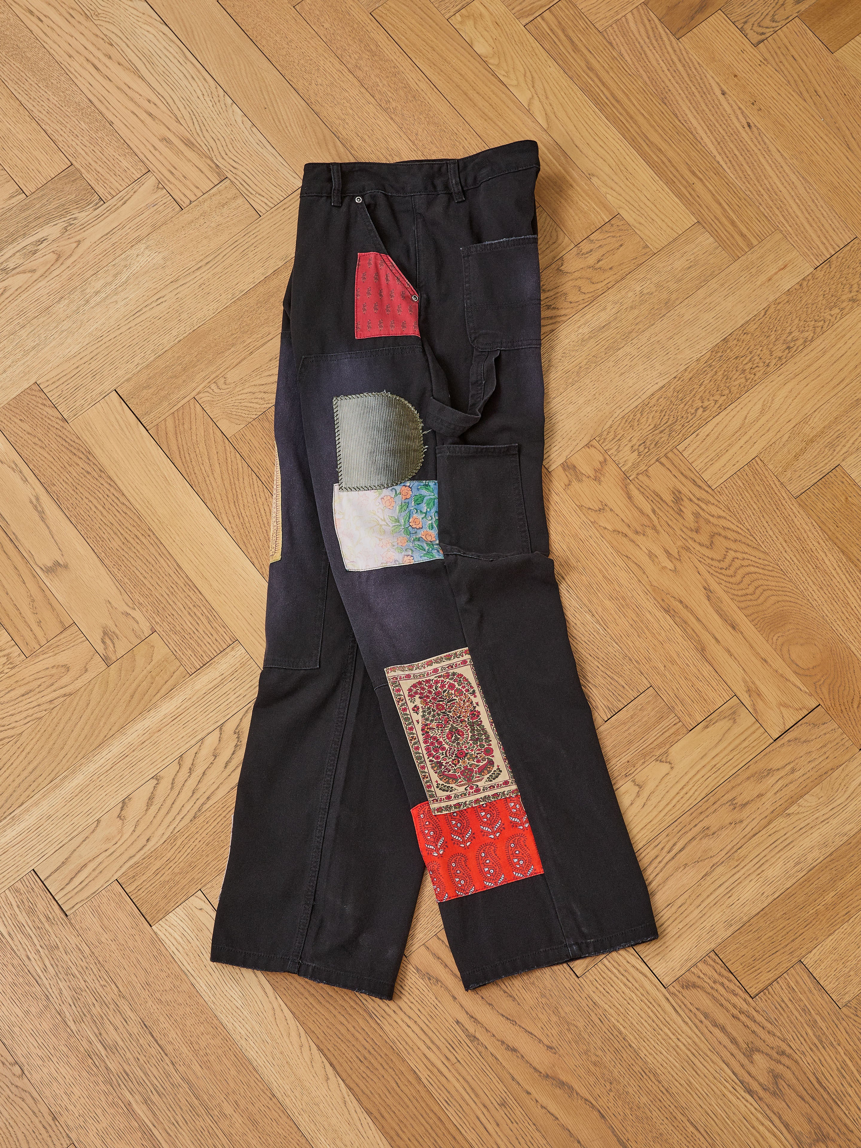 FOUND’s Tapestry Carpenter Canvas Pants—black twill with colorful fabric patches inspired by Pakistani quilts—are laid flat on a light wood herringbone floor.