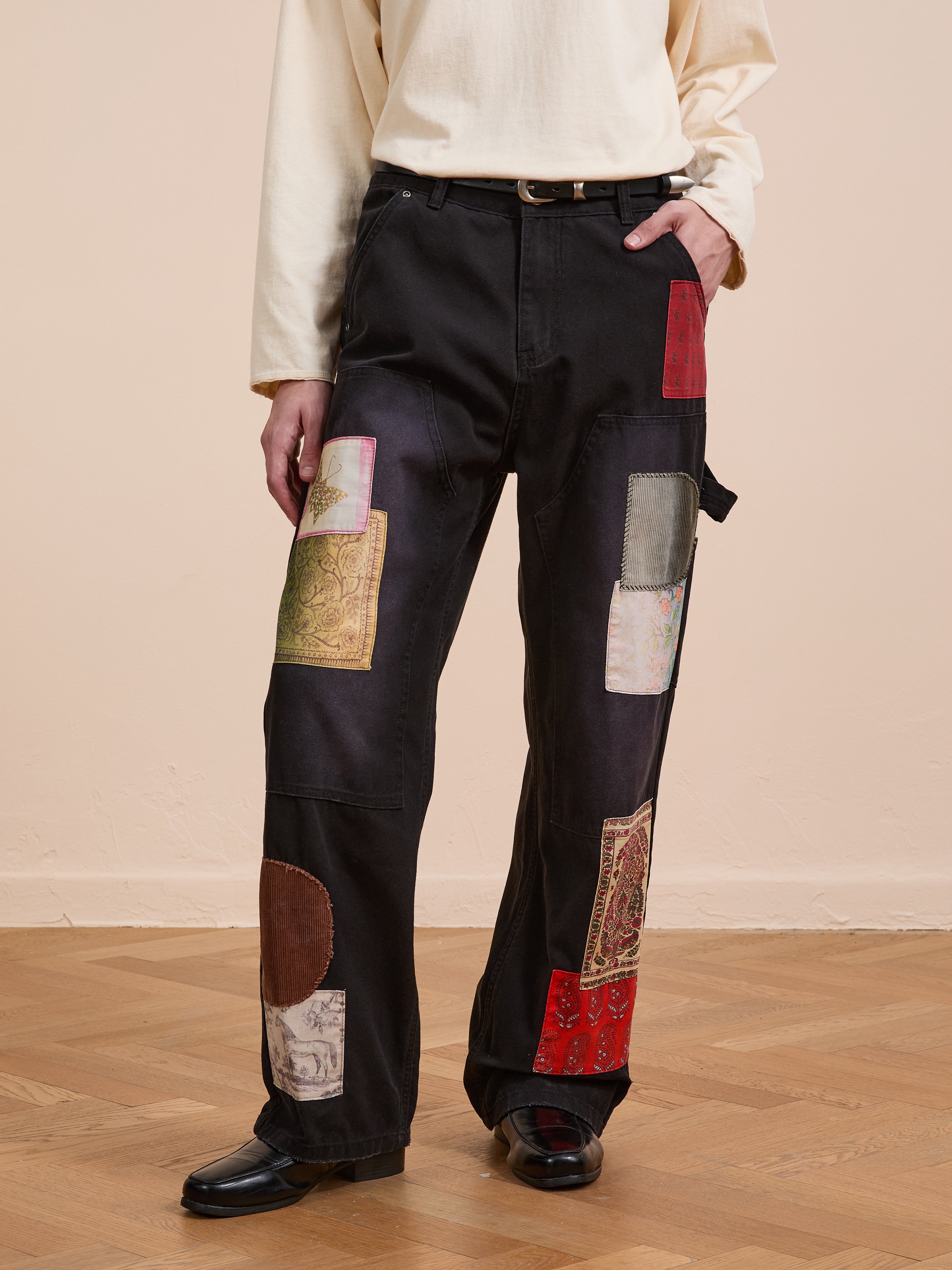 A person wears FOUND's Tapestry Carpenter Canvas Pants with colorful Pakistani quilt-inspired patches, a cream long-sleeve shirt, and black shoes, standing on a wooden floor against a plain beige wall.