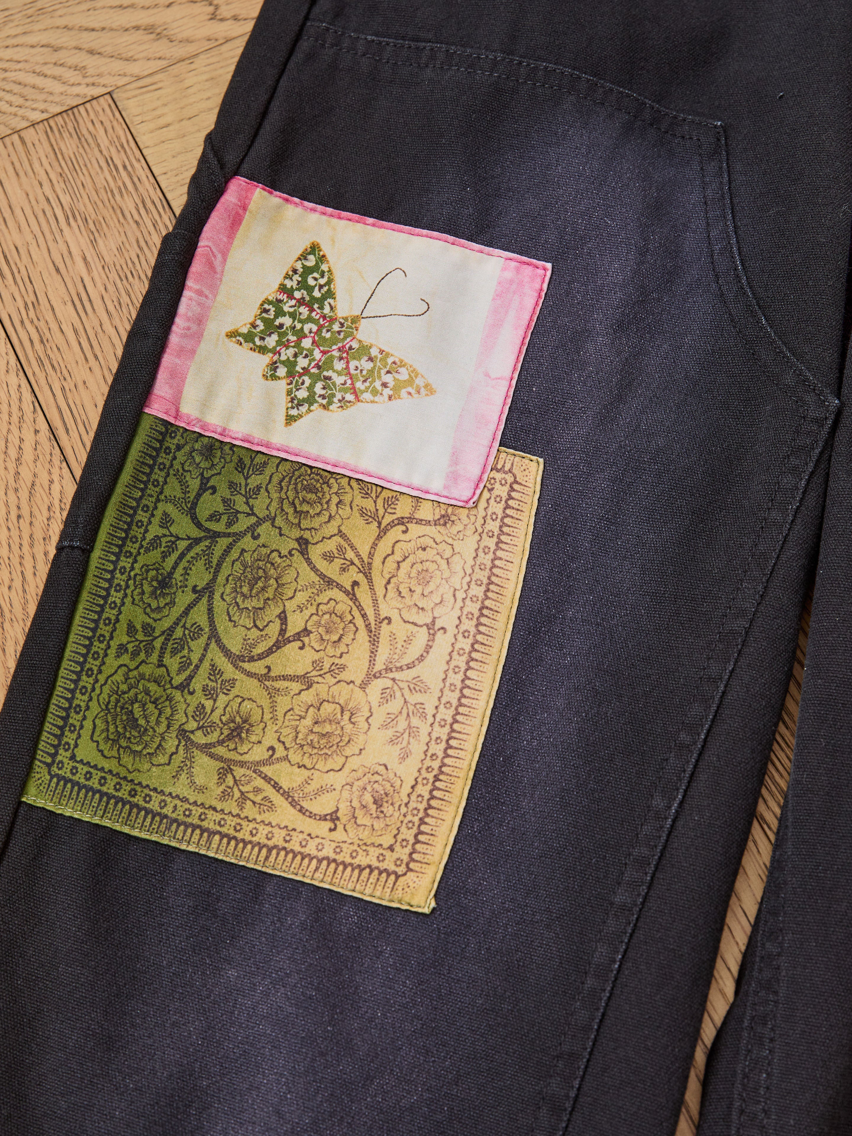 Close-up of FOUND Tapestry Carpenter Canvas Pants—unisex relaxed fit with two decorative patches: a floral butterfly and an ornate floral motif on yellow-green, inspired by Pakistani quilt designs.