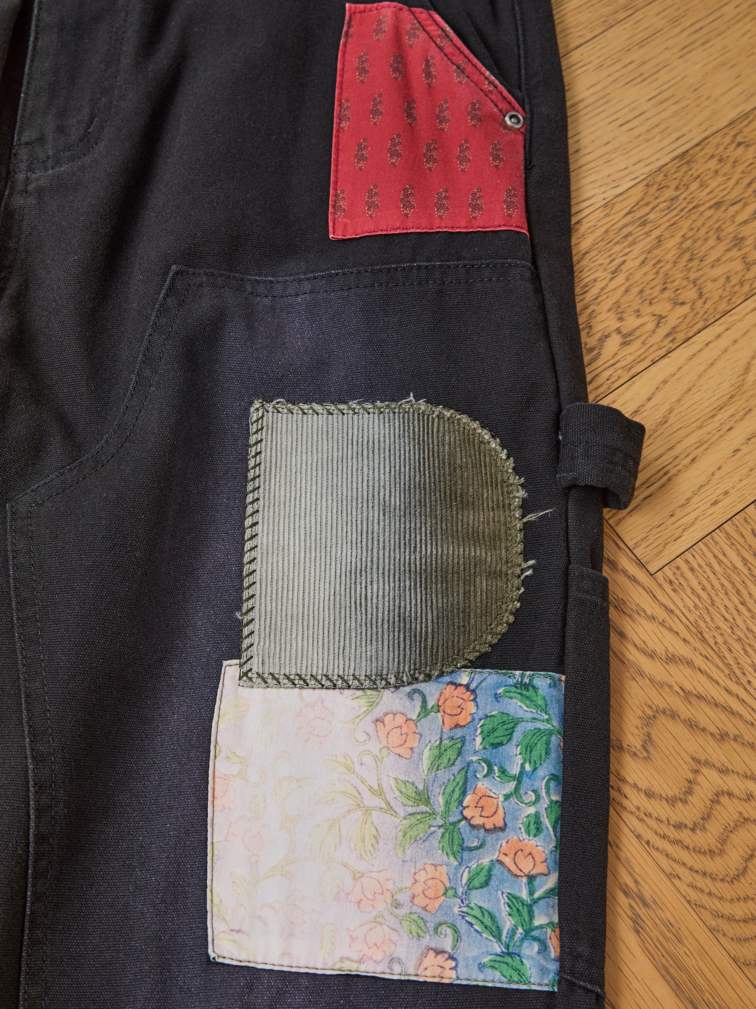 Close-up of FOUND Tapestry Carpenter Canvas Pants featuring black twill with red patterned, green corduroy, and floral fabric patches, highlighting artisan craft, displayed on a wooden floor.