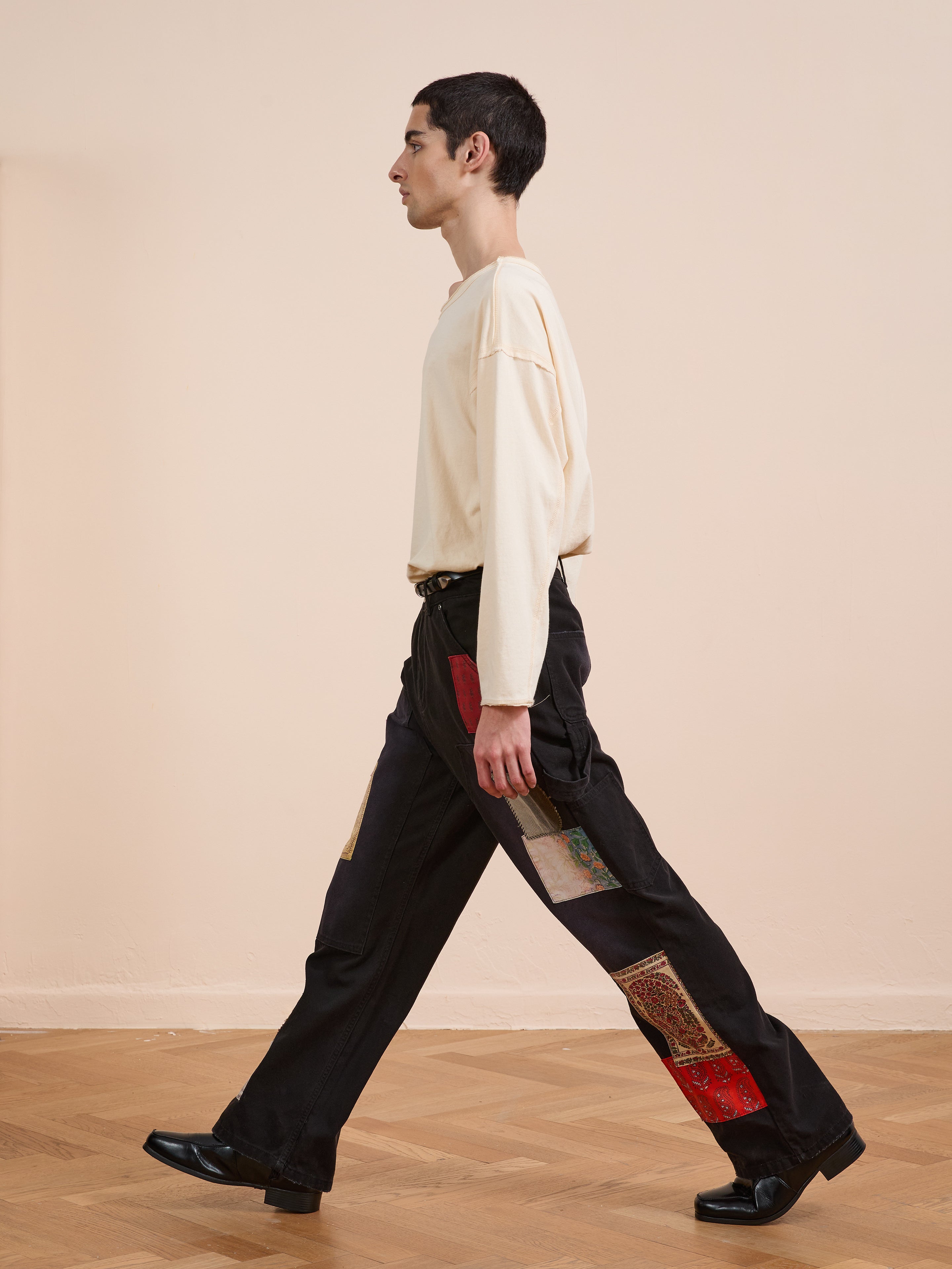A person wears FOUND's Tapestry Carpenter Canvas Pants, featuring colorful patchwork, paired with a cream long-sleeve shirt while walking indoors on a parquet floor against a light wall.