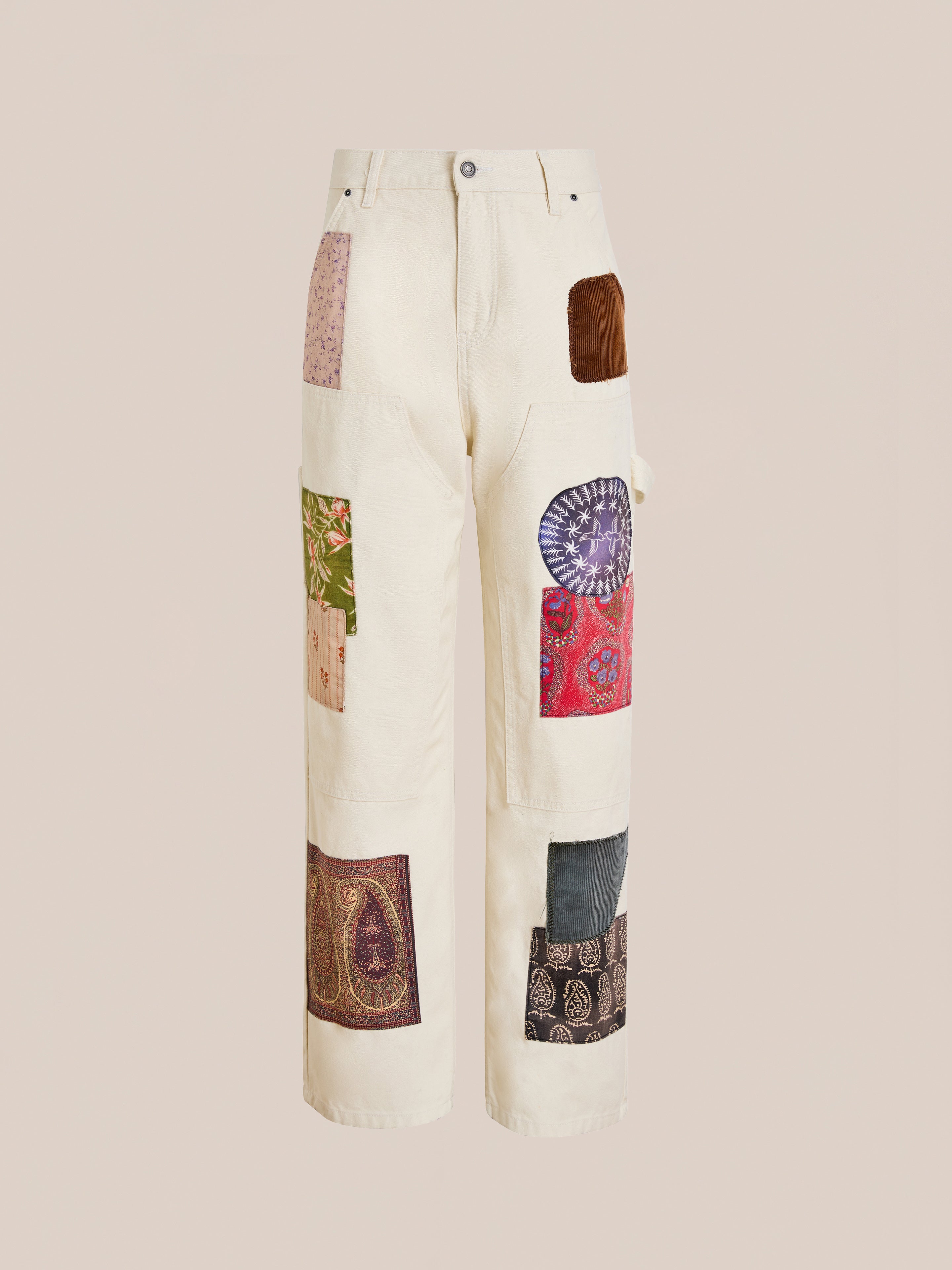 FOUND’s Tapestry Carpenter Canvas Pants are cream-colored unisex relaxed fit pants with multicolored fabric patches inspired by Pakistani quilt designs, sewn on the front and sides. Shown against a plain background.
