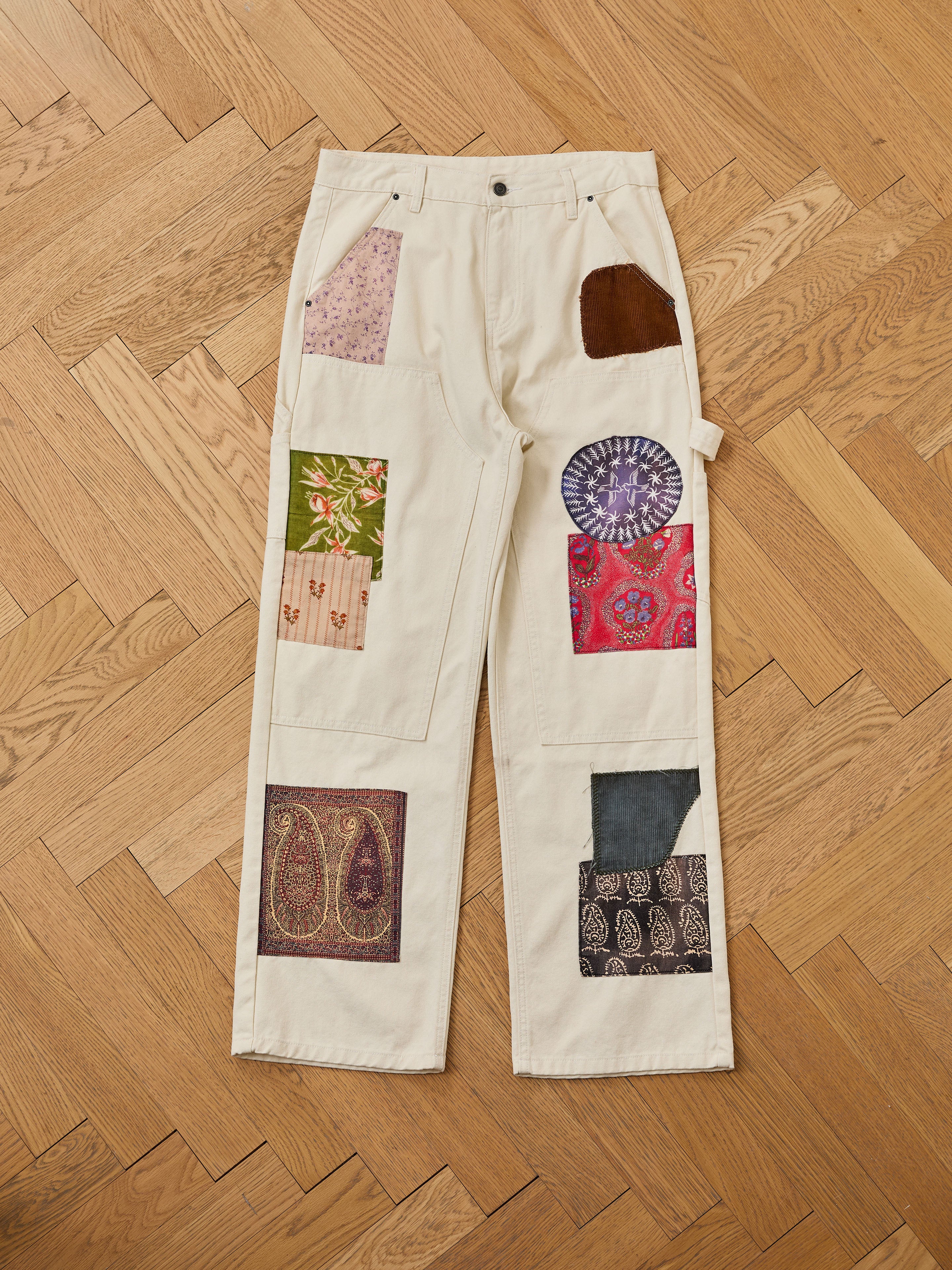 FOUND Tapestry Carpenter Canvas Pants: beige unisex relaxed-fit pants with colorful fabric patches inspired by Pakistani quilt designs, shown laid flat on a wooden herringbone floor.