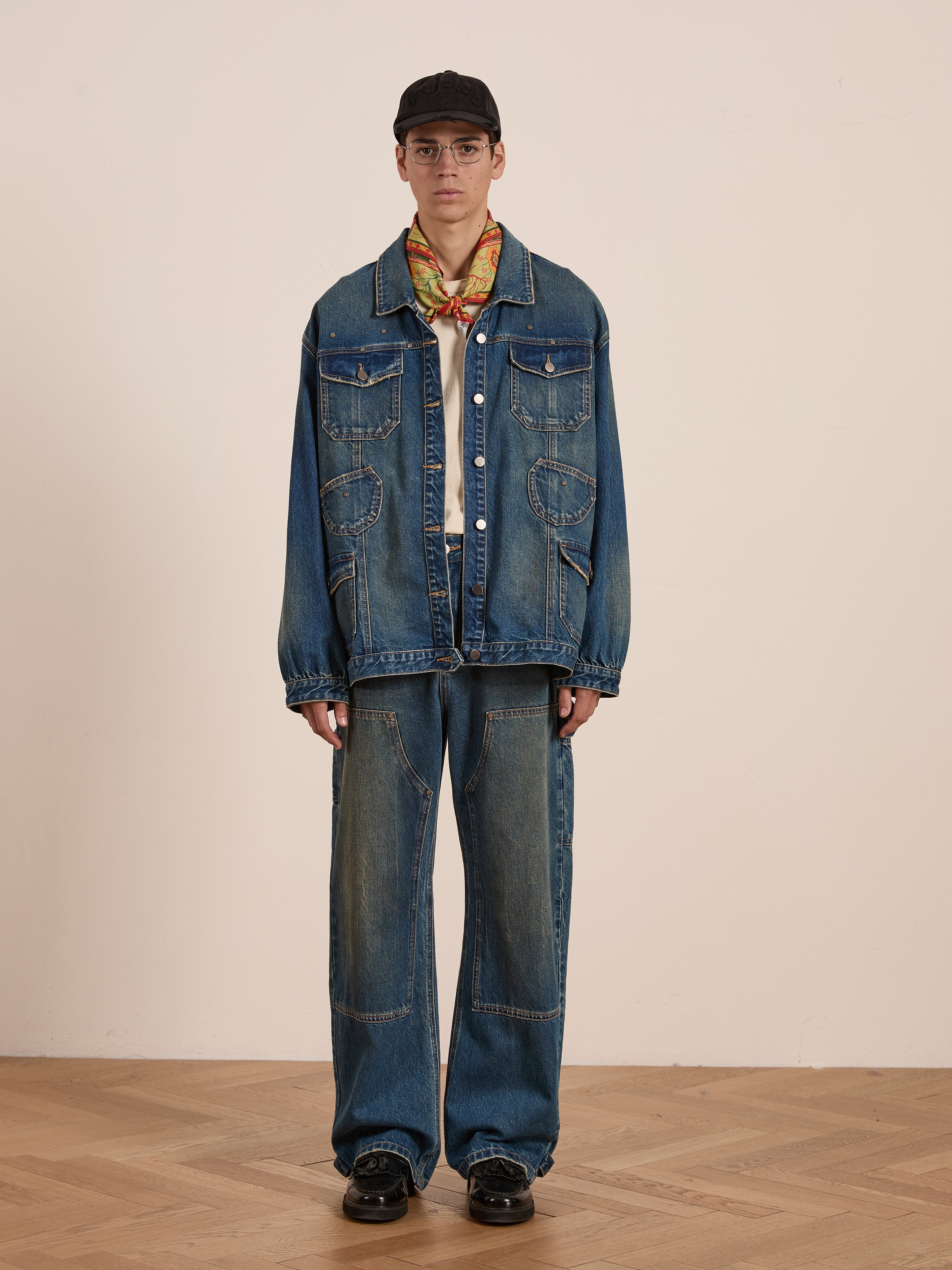 A person stands on a wooden floor against a plain background, wearing FOUND's oversized Denim Carpenter Jacket and jeans, paired with a beige shirt, colorful scarf, black shoes, and cap. This unisex relaxed fit outfit is the ultimate workwear staple.