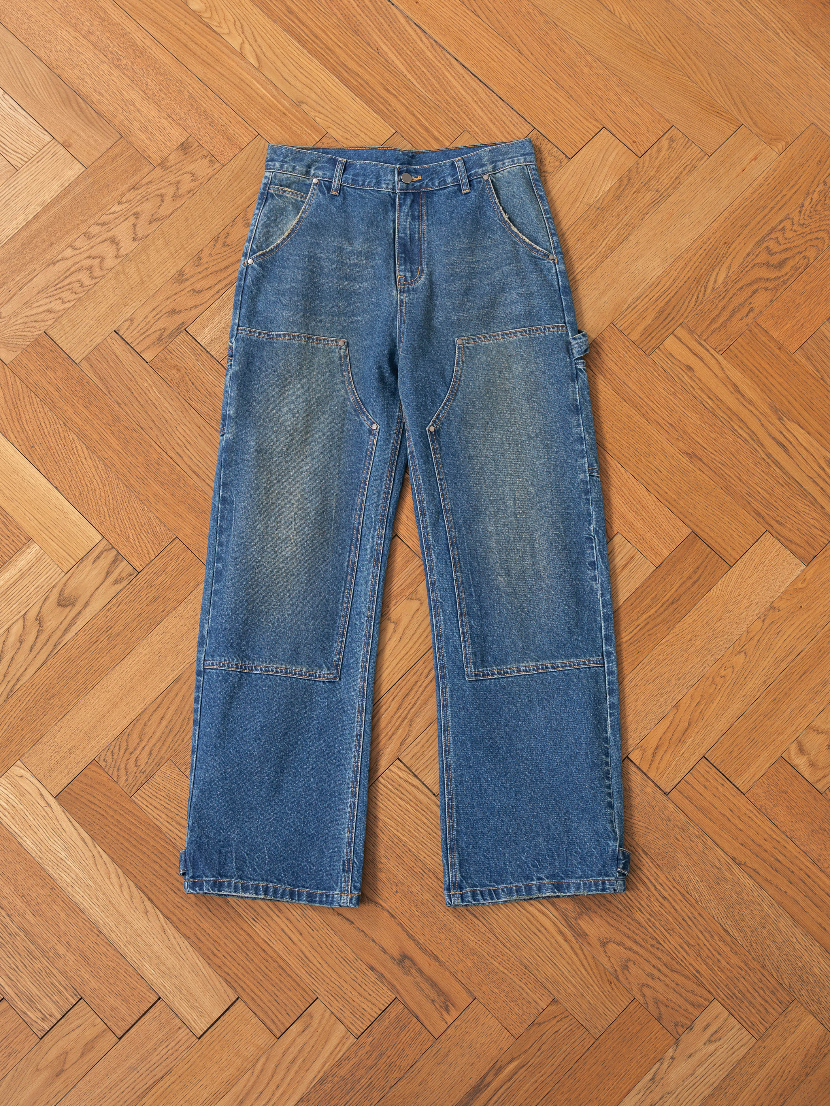 FOUND's Denim Carpenter Jeans feature a vintage-inspired, relaxed straight-leg fit with visible front and knee patches, showcased laid flat on a wooden herringbone floor.