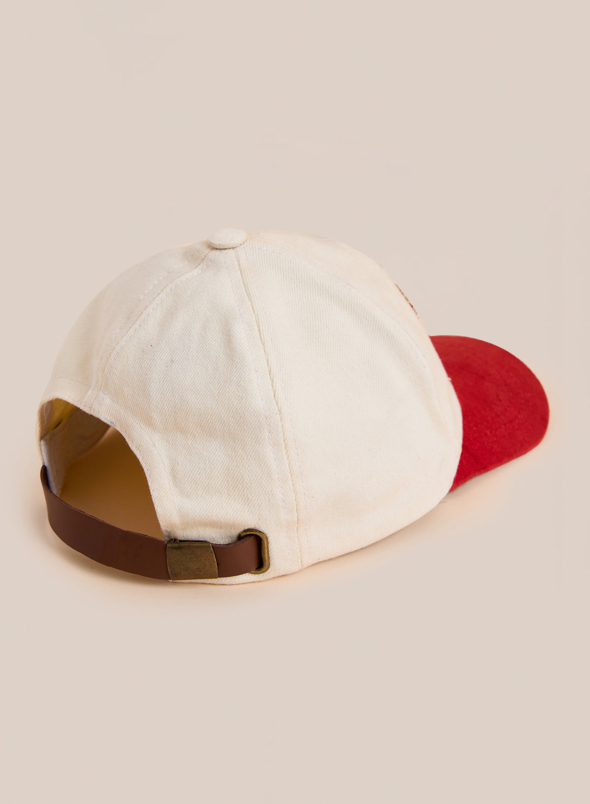 The Club Patch Cap by Found is a beige vintage baseball cap with a red brim, shown from the back with an adjustable brown strap and brass buckle.